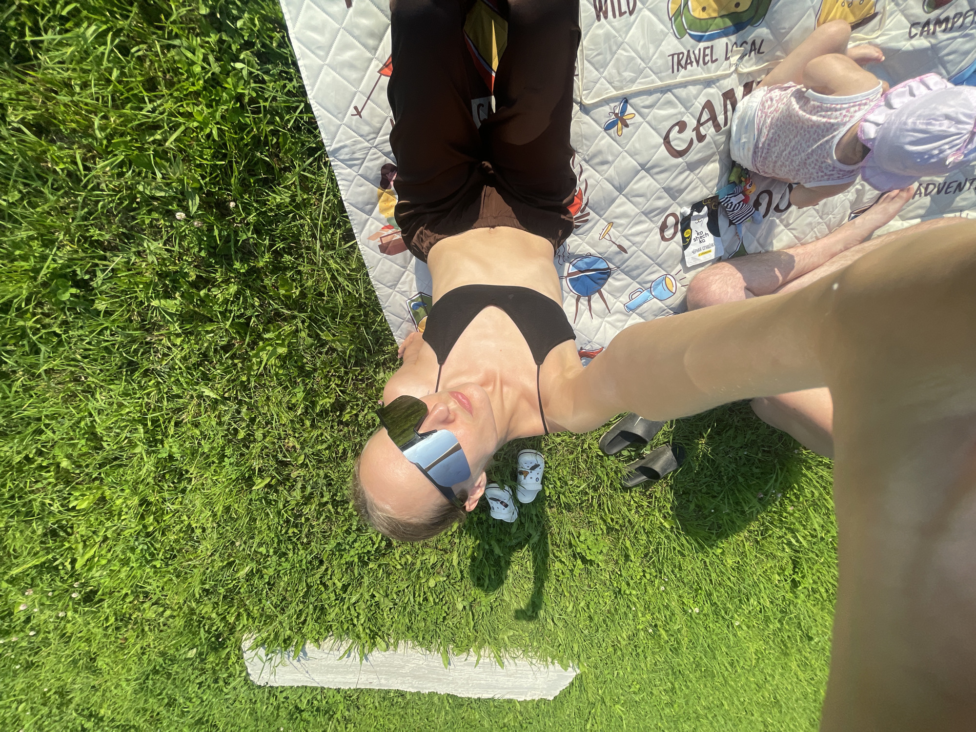 A high-angle shot captures a sunny day scene on a grassy lawn, with a picnic blanket spread out. The blanket is adorned with playful graphics, including depictions of camping gear and animals, with the text "TRAVEL LOCAL" and "CAMPING ADVENTURES" visible.
Two people are present. In the foreground, a person lies on their back, wearing a black bikini top and dark pants. They have on large, mirrored sunglasses and appear relaxed. Near them are a pair of small, blue sandals with cartoon characters. To the right and slightly above, a baby is lying on their back, wearing a pink and white patterned outfit and a light purple hat. The baby's legs are bent, and they are facing away from the camera.
The scene suggests a leisurely outdoor activity, likely a picnic or a day at the park, on a bright, clear day. The bright sunlight casts distinct shadows, indicating it's likely midday. The lush green grass and clear sky suggest a warm weather setting. The overall mood is one of relaxation and enjoyment. There are also two black slides visible on the grass near the adult. A small white pamphlet or tag is visible on the blanket near the baby, with Russian text on it, possibly "кошечк."
