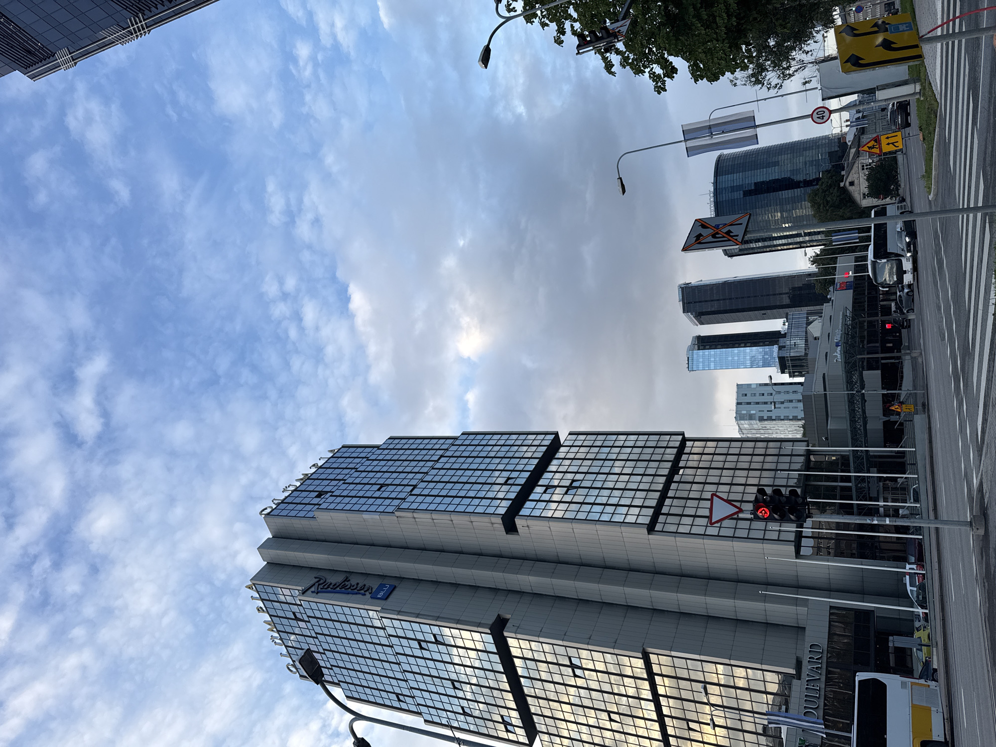 The image depicts a street view in Tallinn, Estonia, dominated by the Radisson Blu Sky Hotel, a prominent skyscraper with a distinctive blue "Radisson" logo. The hotel's facade is made of glass and concrete, reflecting the overcast sky. In the foreground, a traffic light displays a red arrow indicating a no-turn-left signal, while a white triangular yield sign is positioned above it.
Several vehicles are visible on the road, including a white and yellow bus, cars, and a truck. The street is paved and features a pedestrian crossing. A lamppost stands near the traffic light, and another lamppost is visible further down the street.
In the background, other modern buildings, including high-rise skyscrapers, contribute to the urban skyline. Some of these buildings are also made of glass, reflecting the surrounding environment. Trees and foliage are present on the right side of the image, adding a touch of nature to the urban landscape.
The sky is cloudy, with patches of blue sky visible. The overall impression is a bustling city environment with a mix of modern architecture and functional urban infrastructure. The image appears to have been taken during daylight hours.