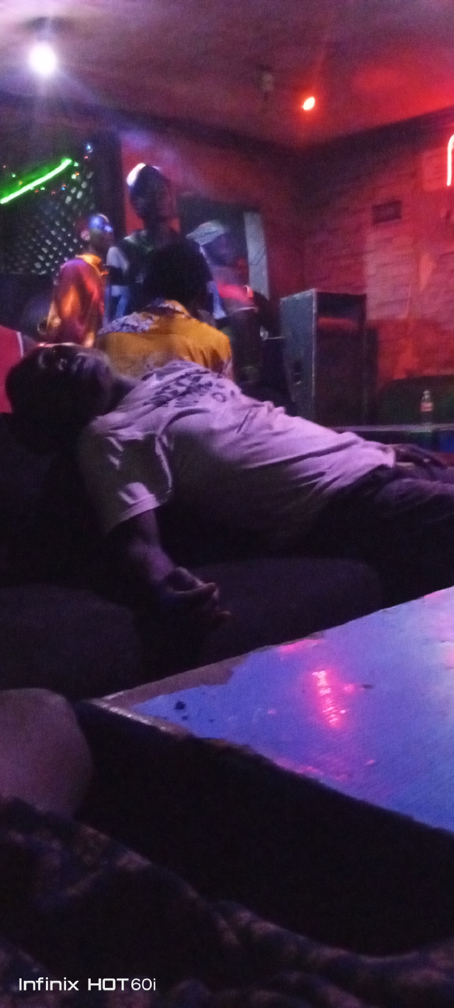 The scene is inside a dimly lit bar or nightclub in Adankolo, Nigeria. A man is lying down on a couch or seating area, appearing to be asleep or passed out. Several other people are standing in the background, possibly performing on a stage or gathering near a designated area. There is a table in the foreground with a shiny surface. The environment is characterized by low lighting, with various colored lights illuminating parts of the scene. A speaker is visible behind the sleeping man.