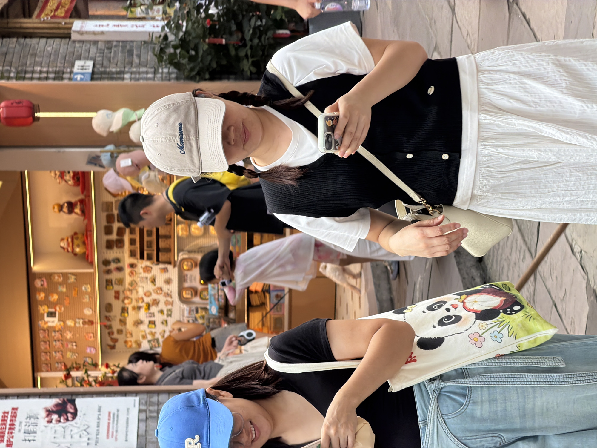 In Yuzhong District, China, a street scene unfolds with two women in the foreground. The woman on the left, partially visible, wears a blue baseball cap, glasses, and a black t-shirt. She carries a white tote bag adorned with a cartoon panda graphic. To her right, a woman wearing a beige baseball cap with "Mamasma" embroidered in blue, and a black vest over a white t-shirt, looks down at her smartphone. Her hair is styled in two braids, and she carries a beige cross-body bag. In the background, a shop displays various items on shelves, including figurines and what appear to be stickers. Other pedestrians are visible in the background, moving through the bustling street. The environment is characterized by brick and stone architectural elements.