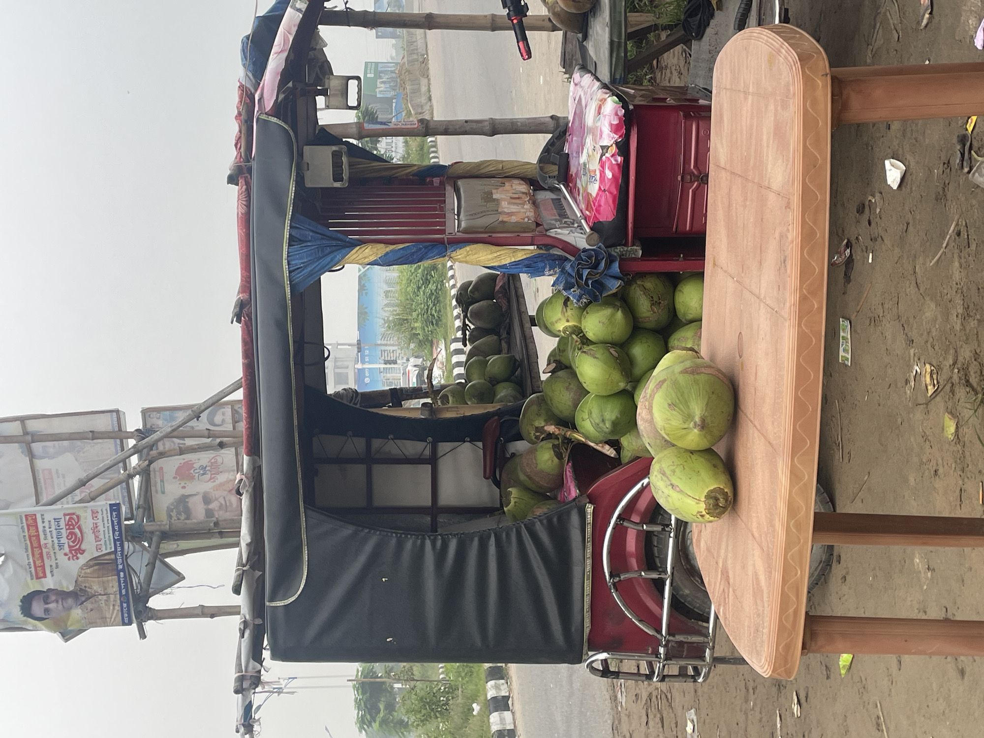Here's a concise description of the image:
The image shows a coconut vendor's rickshaw in what appears to be Purbachal, Bangladesh. The rickshaw is loaded with fresh green coconuts. A makeshift table is attached to the rickshaw, holding additional coconuts. The rickshaw has a black weather shield. Political posters are attached to a bamboo structure on the rickshaw. The environment suggests an urban or suburban setting with a road.