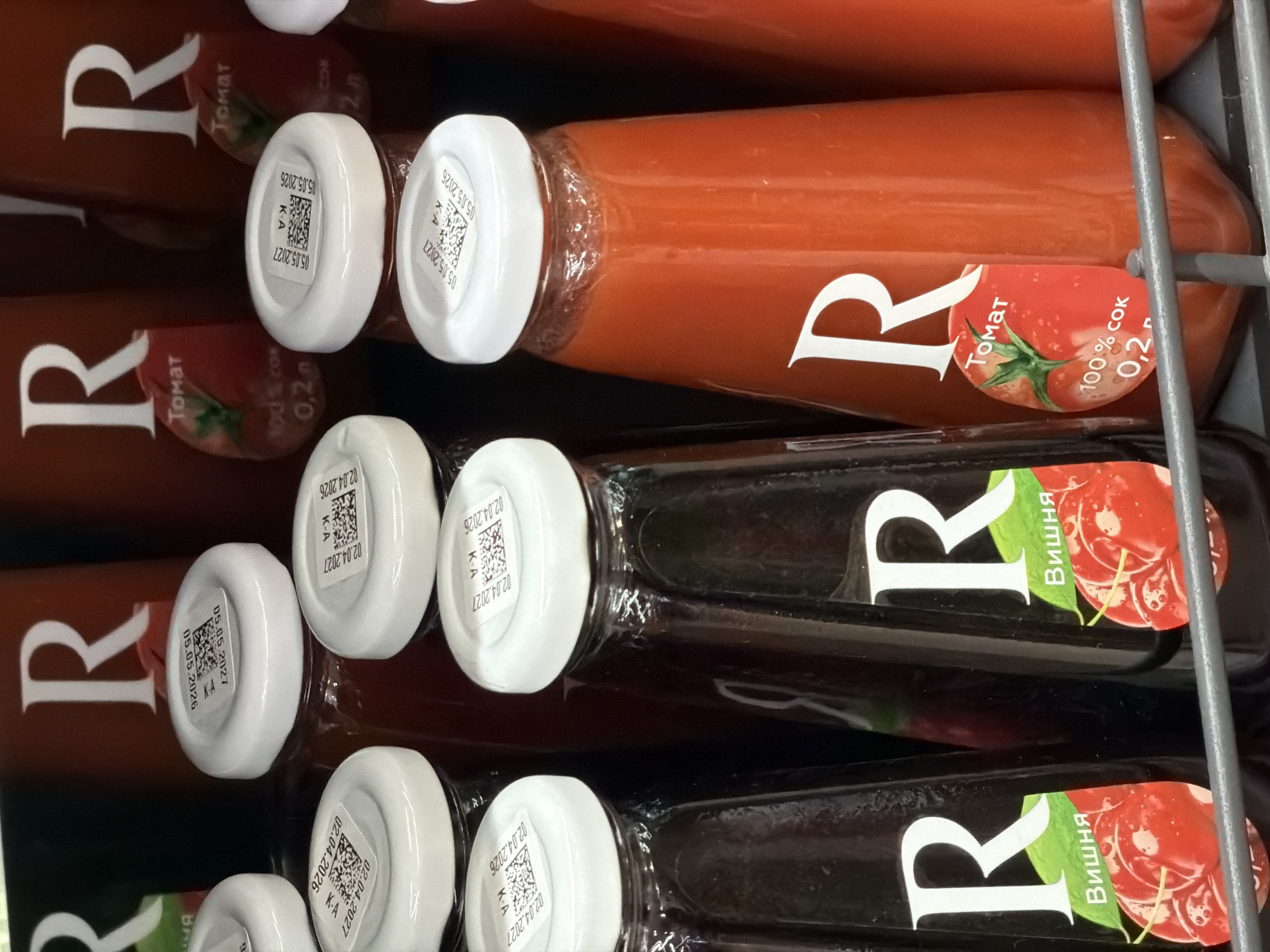 The image shows a shelf in a store, likely a supermarket or convenience store, displaying several bottles of juice. The bottles are dark glass and have white caps. The labels on the bottles are predominantly white with a large "R" logo. Some bottles are labeled "Вишня" (Cherry) and feature an image of cherries. Other bottles are labeled "Томат" (Tomato) and have an image of a tomato. The lighting appears to be artificial, casting reflections on the glass bottles. The bottles are arranged in rows on a metal shelf. The background is dark and out of focus, but suggests the presence of more products or shelving. There are no people visible in the image. The presence of Russian or Belarusian language on the labels, such as "Томат" and "Вишня," combined with the typical presentation of products in a retail environment, suggests this scene could be located in Minsk, Belarus, a city where such products are commonly sold. The specific location within Minsk cannot be determined solely from the visual information.