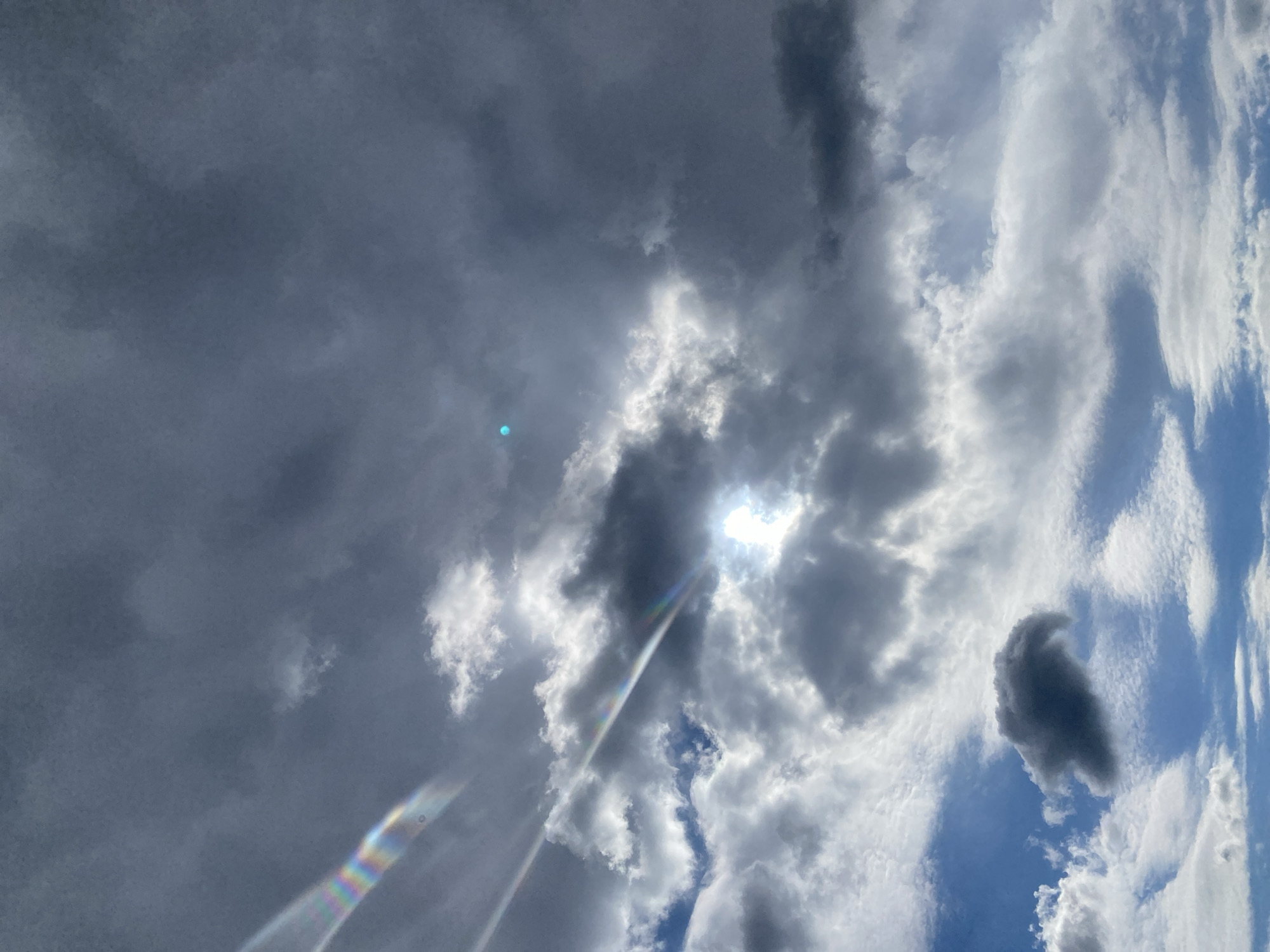 The image displays a dynamic, upward-facing view of a cloudy sky during daytime. The scene is dominated by various cloud formations, ranging from dense, dark grey clouds occupying the left and central portions to brighter, more ethereal white and wispy clouds towards the right. Patches of clear blue sky are visible between the clouds, particularly on the right side of the frame.
The sun, though obscured by clouds, is the central source of light, creating a brilliant burst of radiance from behind a cluster of bright, white clouds near the image's center. This intense light illuminates the edges of the surrounding darker clouds, creating a dramatic interplay of light and shadow and highlighting the textures within the cloudscape.
Two distinct lens flares are visible, indicating the sun's direct interaction with the camera lens. A prominent, elongated flare exhibiting a rainbow spectrum extends diagonally from the lower-left to the upper-middle of the image. Another smaller, circular blue-green flare is located in the upper-left quadrant amidst the darker clouds.
The weather appears partly cloudy, with moments of strong sunlight breaking through. There are no identifiable human subjects, man-made objects, or visible text. The image captures the vastness and ever-changing nature of the sky, with the contrasting light and dark clouds creating a sense of depth and visual interest.