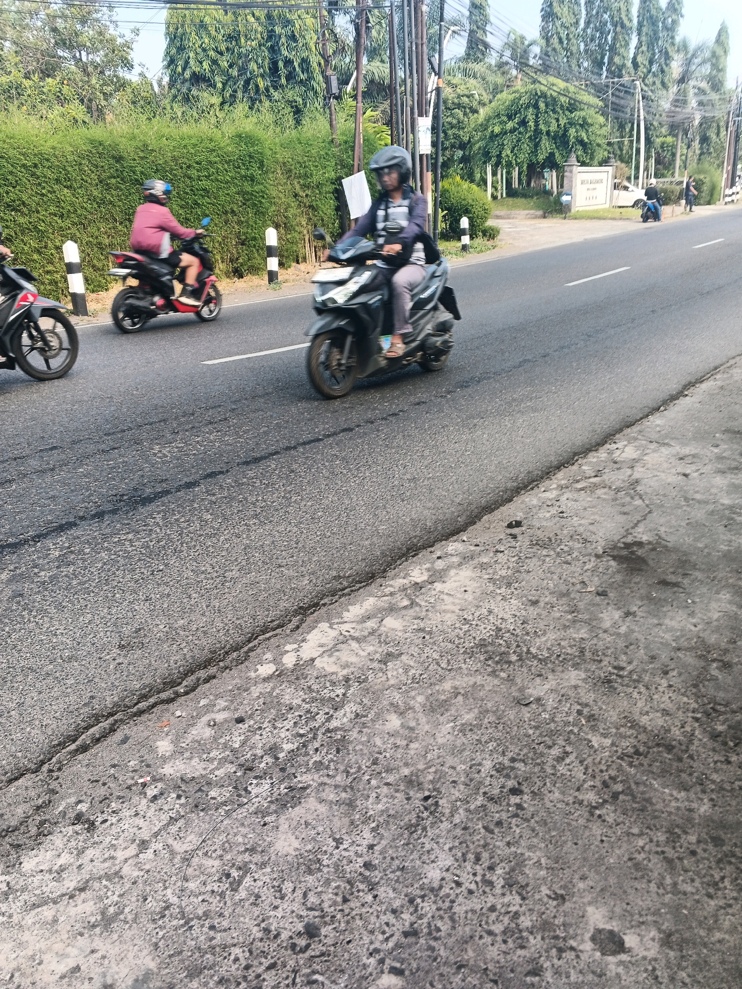 In Ungaran, Indonesia, on a sunny day, a person wearing a helmet rides a black motorcycle in the middle lane of a two-lane asphalt road. The rider is wearing a striped long-sleeved shirt and grey pants, and their feet are visible on the footpegs. Behind them, a person in a pink jacket and helmet is also riding a motorcycle, which is red. The road is lined with green hedges on the left side and trees on the right. Further down the road, a sign reads "WIRA MANDIRI". The foreground of the image shows the rough texture of the asphalt and a concrete edge.