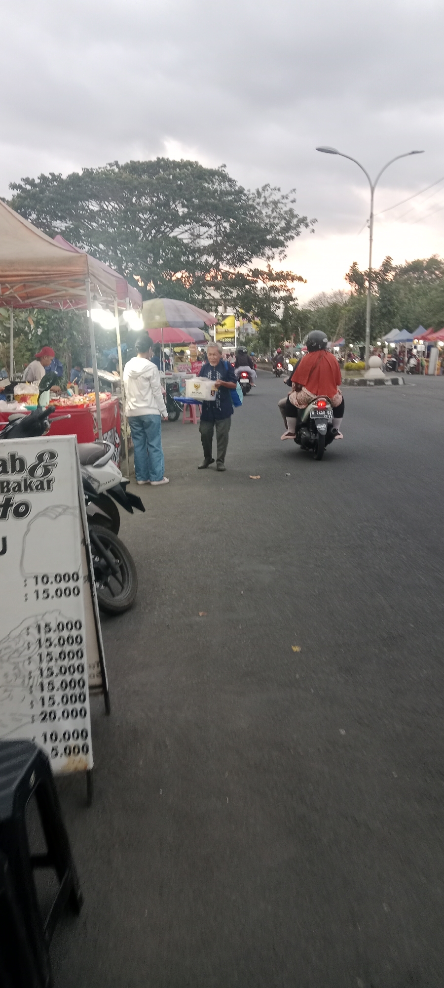 The image depicts a street scene in Ngemplak, Indonesia, likely a market area, given the presence of stalls and vendors. In the foreground, a white sign with black text, partially obscured by a motorcycle, lists prices such as "10.000" and "15.000". To the left, a food stall is visible with a red awning and various food items displayed. A person wearing a red cap is tending to the stall.
Further into the scene, a woman in a white hoodie and blue jeans stands near a food stall, facing away from the camera. To her right, an elderly person in a blue shirt and green pants is holding a white box and appears to be walking. A motorcycle with a rider wearing a red hijab is in motion on the right side of the road, heading away from the camera. Another motorcycle is parked in the foreground.
The background shows more market stalls, trees, streetlights, and a cloudy sky. The overall impression is a busy outdoor market setting in the late afternoon or early evening.