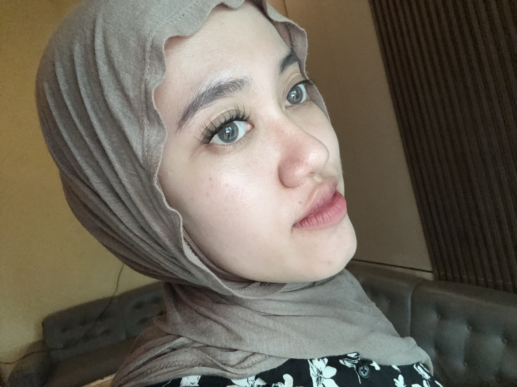 A young woman wearing a taupe-colored hijab is shown from the chest up. The lighting is soft and diffused. The woman appears to be in a residential interior. She has light skin, prominent cheekbones, full lips, and greyish-blue eyes. Her eyebrows are defined, and she is wearing false eyelashes. She is looking slightly upward. Part of her black and white floral-patterned top is visible in the lower part of the frame. The background includes a wall of light brown and a dark, tufted sofa. The location is Gadingrejo, Indonesia.