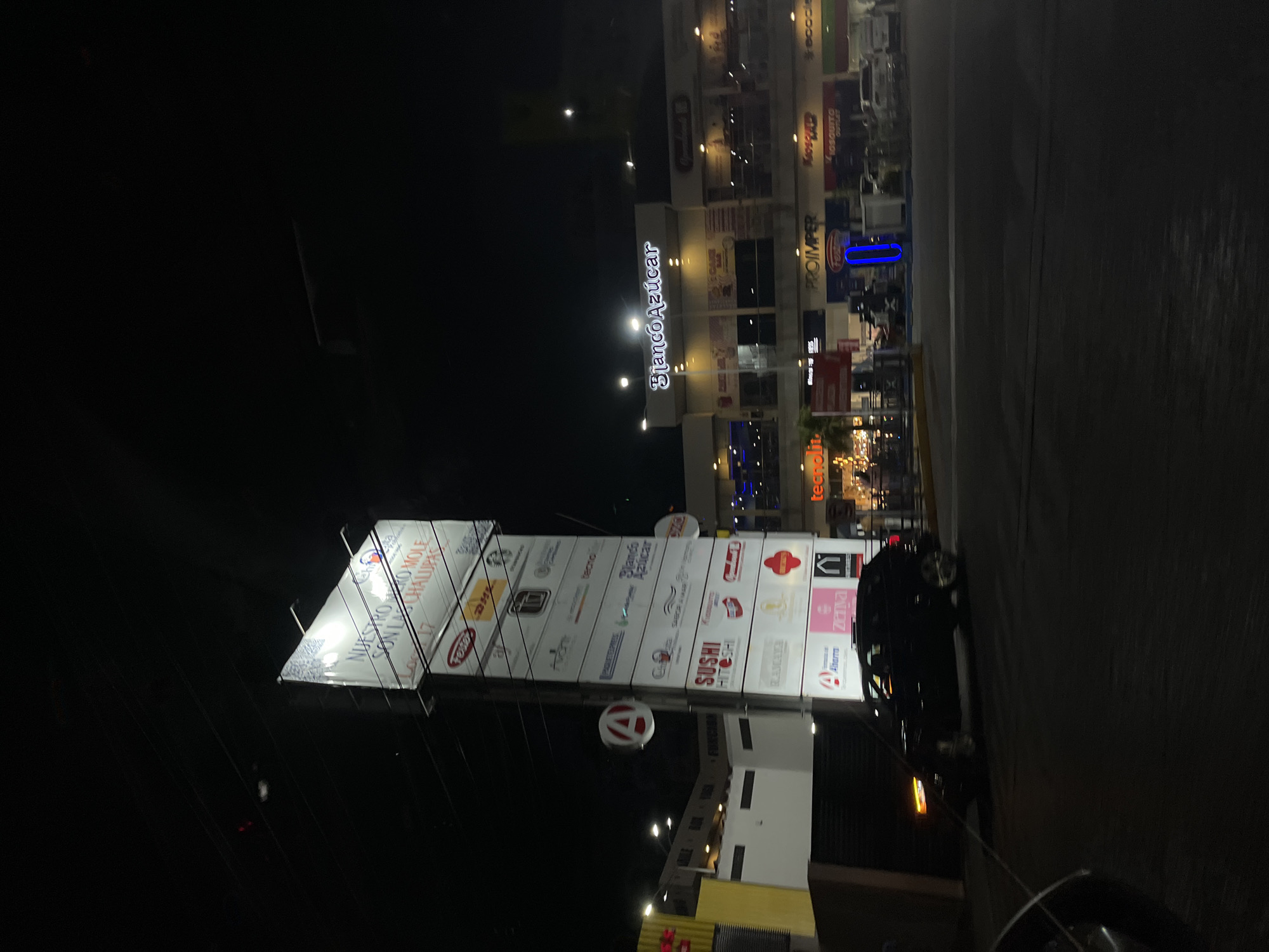 The image shows a street scene in San Bernardino Tlaxcalancingo, Mexico, at night. A large sign displays logos of businesses in a nearby shopping plaza. These include "Nuestro Mole," "DHL," "Blanco Azúcar," "Sushi Hitoshi," "Tecnolit," "Proimper," "Mosquito Outlet," and "Ecocle." A shopping center with the words "Blanco Azúcar" is visible in the background, with multiple levels and illuminated windows. A car is parked near the sign and the foreground is dark. Overhead power lines crisscross the sky. The scene is dimly lit by streetlights and building lights.