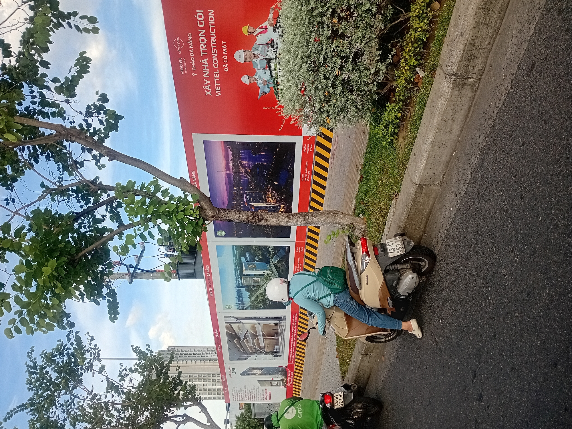 The image depicts a street scene in Da Nang, Vietnam, with a focus on a large construction hoarding. The hoarding is red and white, featuring promotional images of modern buildings and text in Vietnamese. The text on the right side of the hoarding includes "VIETTEL aiHomes CONSTRUCTION", "CHÀO ĐÀ NẴNG", "XÂY NHÀ TRỌN GÓI VIETTEL CONSTRUCTION ĐÃ CÓ MẶT", and illustrations of construction workers. On the left side, there are images of architectural designs and text related to "LEED".
In the foreground, two individuals on motorbikes are visible. The person in the foreground is riding a beige scooter, wearing a light blue shirt, blue jeans, a white helmet, and carrying a green backpack. The other person, further back on the left, is on a dark motorbike, wearing a green jacket with a "Grab" logo and a dark helmet. The street is paved, with a concrete curb and a grassy area beyond. Trees with green foliage are present, and in the background, a tall, modern building is visible, along with utility poles and electrical equipment. The sky is partly cloudy and blue.