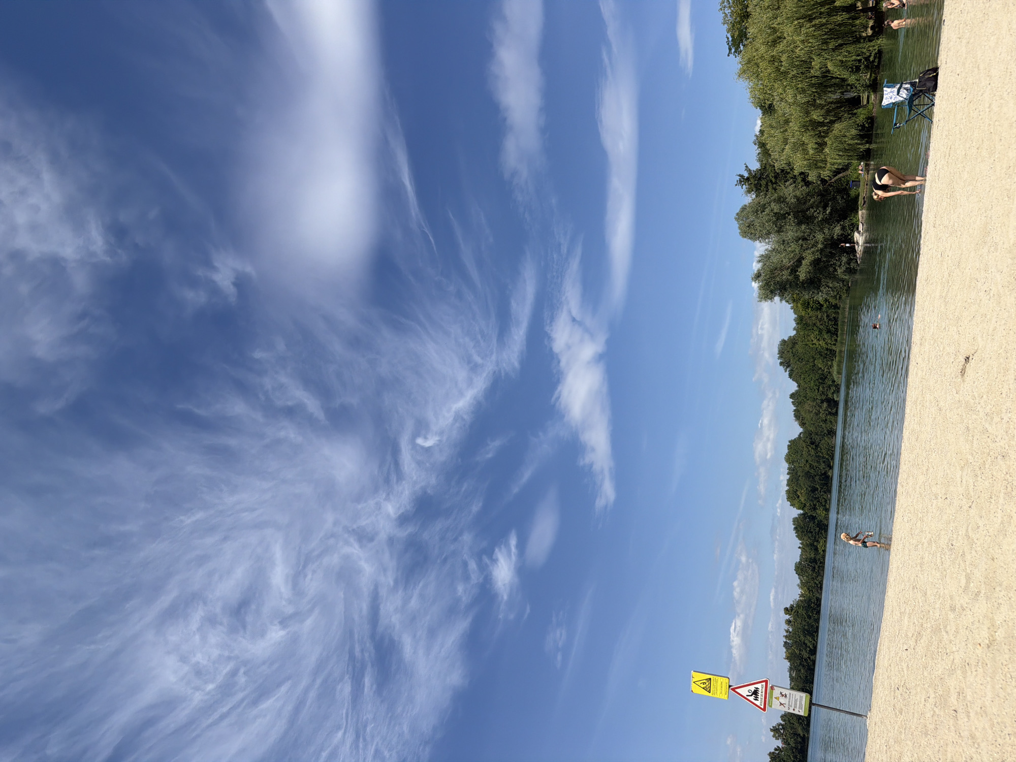 This media file depicts a sunny day at a beach area by a body of water, likely a river or lake, in Strasbourg, France. The scene is dominated by a bright blue sky with wispy white clouds stretching across the frame. The foreground is a sandy beach, leading to the water's edge.
Several people are visible enjoying the day. On the beach, a person is bent over near the water. Further out in the water, a person is wading, and another individual is visible swimming in the distance. Along the treeline bordering the water, some people appear to be relaxing. Trees form a dense green backdrop to the water.
Two warning signs are prominently displayed on a post near the beach. The first sign is triangular with a yellow background and a black border, featuring a lightning bolt symbol, indicating a potential electrical hazard or thunderstorm warning. The second sign, rectangular and also yellow with a black border, shows pictograms of people swimming and jumping, with a crossed-out symbol, suggesting a warning related to water activities or safety.
The overall atmosphere appears to be one of leisure and outdoor recreation on a clear, summer day. The lighting suggests it is midday or early afternoon. The presence of beachgoers and the clear sky indicate pleasant weather conditions.
