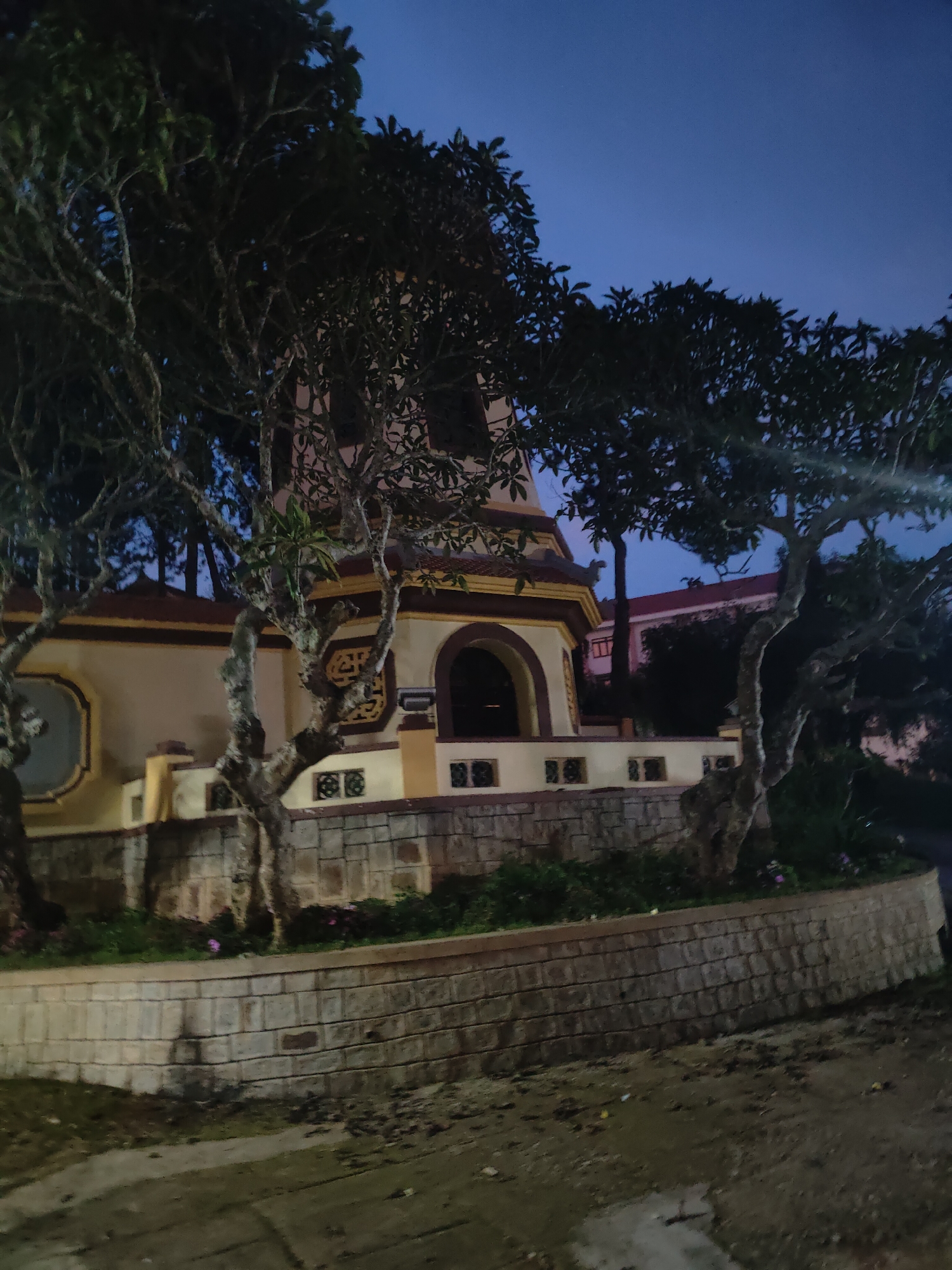 This media file captures a view of a Buddhist stupa at dusk in Da Lat, Vietnam. The sky is a deep, clear blue, indicating the transition from day to night. The stupa, a tall, multi-tiered structure with ornate yellow and brown accents, is partially obscured by the branches of two large trees in the foreground. The trees have gnarled, textured trunks and dark green foliage, their silhouettes stark against the twilight sky.
The stupa sits behind a low, curved wall constructed of light-colored bricks. This wall encloses a small garden area with green bushes and a few scattered purple flowers, adding a touch of color to the scene. The building itself is painted a pale yellow and features arched openings and decorative patterns. In the background, beyond the stupa and trees, a building with a red roof is visible, suggesting an urban or semi-urban setting.
The ground in the foreground is uneven, appearing to be a mix of dirt and possibly some scattered debris, with a small puddle reflecting the dim light. The overall atmosphere is serene and contemplative, fitting for a religious site at the end of the day. The lighting suggests it is either early evening or late dusk, with artificial lights likely illuminating the stupa and its surroundings, though they are not directly visible. No people are present in the image, emphasizing the stillness of the location. No text is visible.