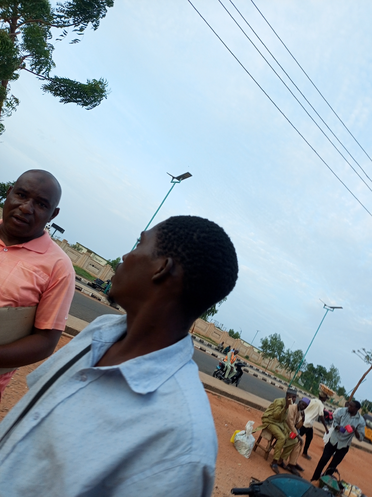 In Gombe, Nigeria, two men stand facing each other on a dirt roadside. The man on the left, wearing a peach polo shirt and holding a cardboard object, looks directly at the camera. The man on the right, facing away from the camera and towards the left, wears a light blue collared shirt. In the background, a road runs horizontally with a sidewalk and a low wall separating it from the street. Several people are visible on the sidewalk and along the road. One person rides a motorcycle, while others stand or sit in small groups. A street lamp with a solar panel is visible in the sky, and power lines stretch across the frame. Trees and foliage line the street in the distance. The sky is overcast with a pale blue hue.