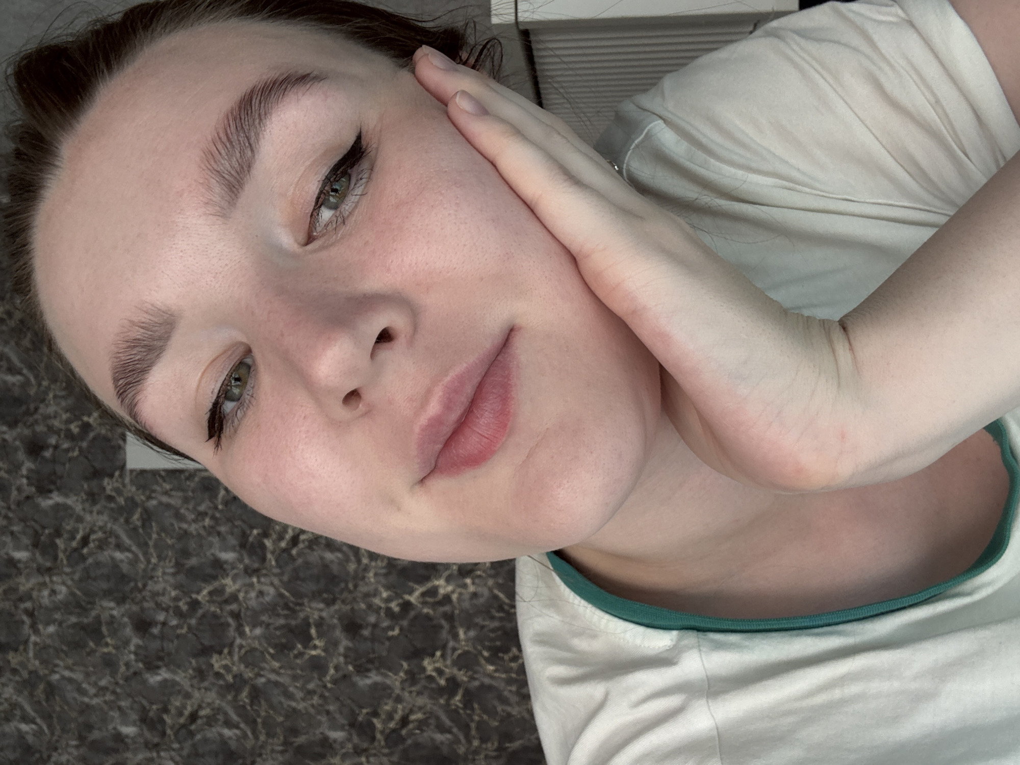 A close-up, angled selfie shows a woman with fair skin, brown hair, and green eyes wearing makeup with winged eyeliner. She's wearing a white t-shirt with a green trim around the neck. Her right hand is touching the right side of her face. The background consists of a dark, marbled surface and what appears to be a white, slatted blind. There is no evidence of a location in the city of Izhevsk, Russia.
