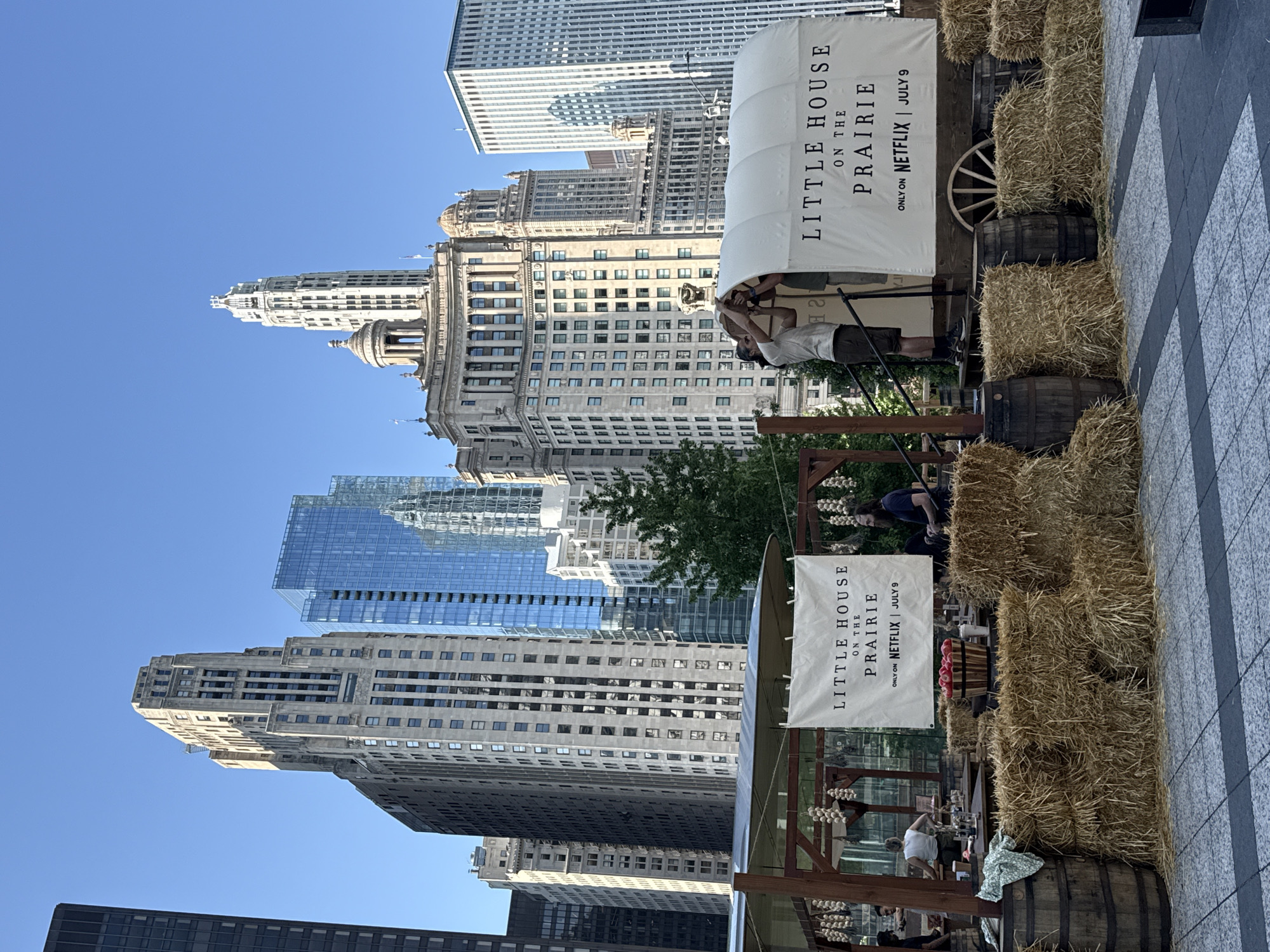 This is a full shot, eye-level, outdoor daytime photograph of a promotional display for "Little House on the Prairie" on Netflix. The display features a white covered wagon with "LITTLE HOUSE ON THE PRAIRIE" printed in large black letters, and "ONLY ON NETFLIX | JULY 9" below. Two individuals are visible behind the wagon, appearing to be adjusting its cover. The wagon is surrounded by bales of hay and wooden barrels. A wooden structure with a roof is to the left, with hanging bunches of garlic or onions. Several people are seen in the background of this structure.
The background is dominated by the Chicago skyline, showcasing a variety of modern and art deco skyscrapers. Notable buildings include tall, rectangular glass towers and older, more ornate structures with distinctive architectural details and setbacks. The sky is clear and blue. The ground in the foreground is paved with large, grey square tiles. The overall impression is a staged event set against the backdrop of a bustling urban environment.