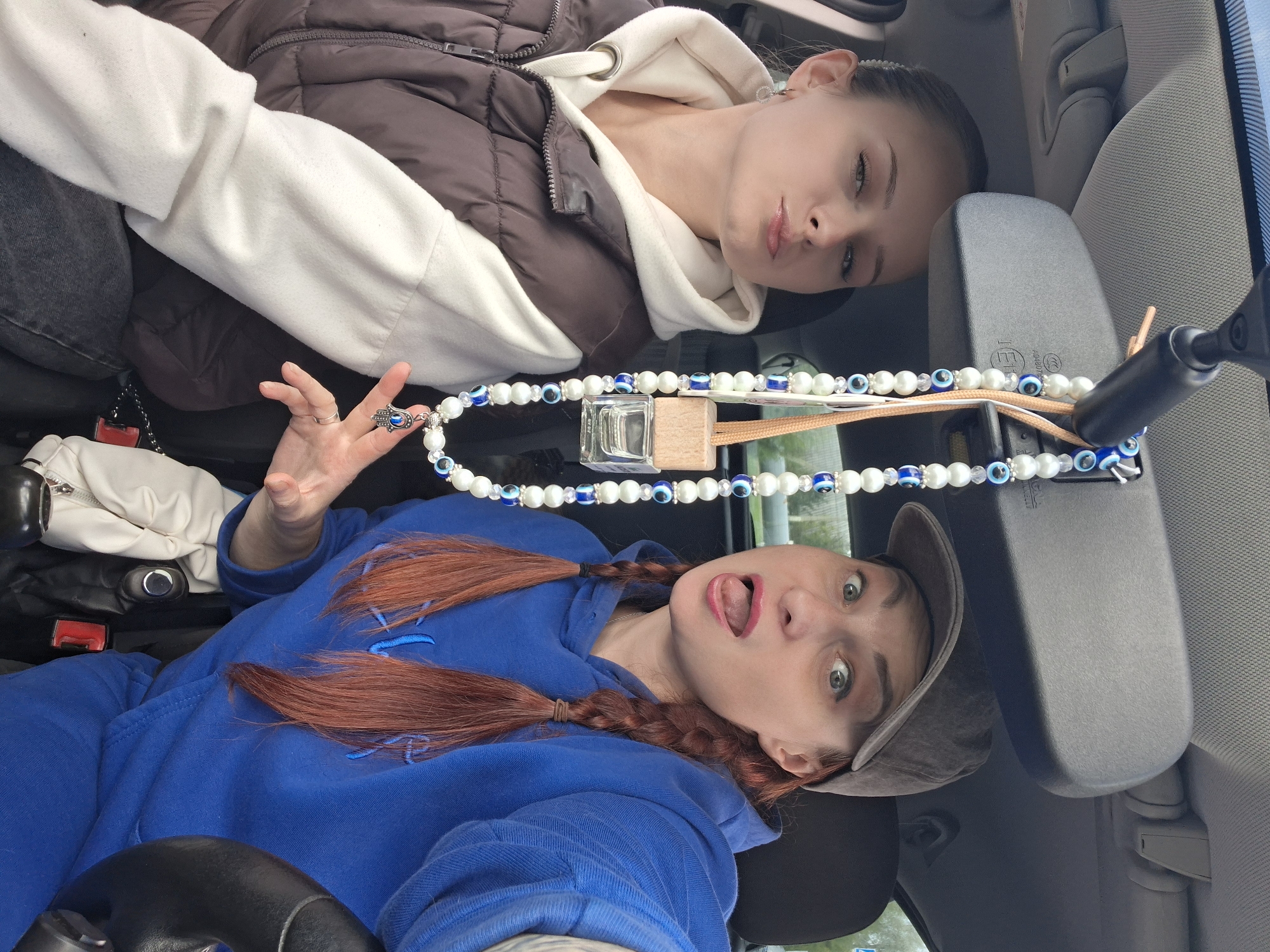 Two young women are inside a car, likely in Myadzyel, Belarus. One woman, with auburn hair in braids, wears a blue hoodie and a gray baseball cap. She is sticking her tongue out at the camera with a playful expression. The other woman, positioned higher in the frame, wears a brown puffer vest over a cream-colored hoodie. She is looking towards the camera with a neutral expression, her head tilted.
A car's rearview mirror is prominently featured, with a string of white and blue evil eye beads hanging from it. A small glass bottle with a wooden cap, possibly an air freshener, is also attached to the mirror. The setting appears to be daytime, with natural light filtering into the car. The car's interior is visible, including seats and a sun visor. There are no discernible weather cues, but the lighting suggests it is not nighttime. The overall mood is casual and lighthearted, with the woman sticking her tongue out conveying a sense of fun. No visible text is present in the image.