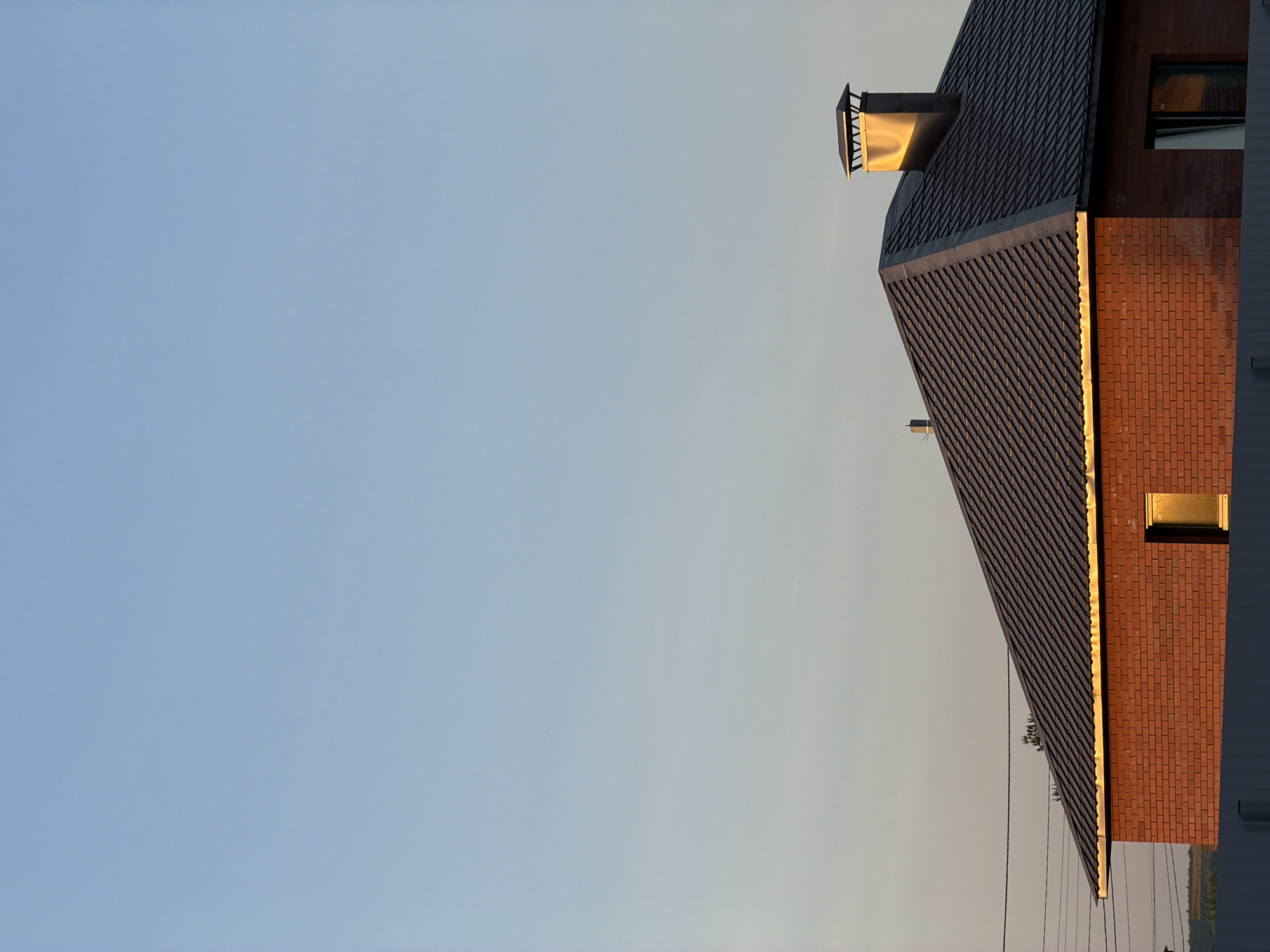 The image depicts the corner of a brick building with a dark roof. The building is likely located in Chaikovsky, Russia, based on the prompt. The building has a chimney with a metal cap. Two windows are visible, reflecting sunlight. Several power lines run across the bottom of the image. The sky is light blue, suggesting it is either early morning or late afternoon.