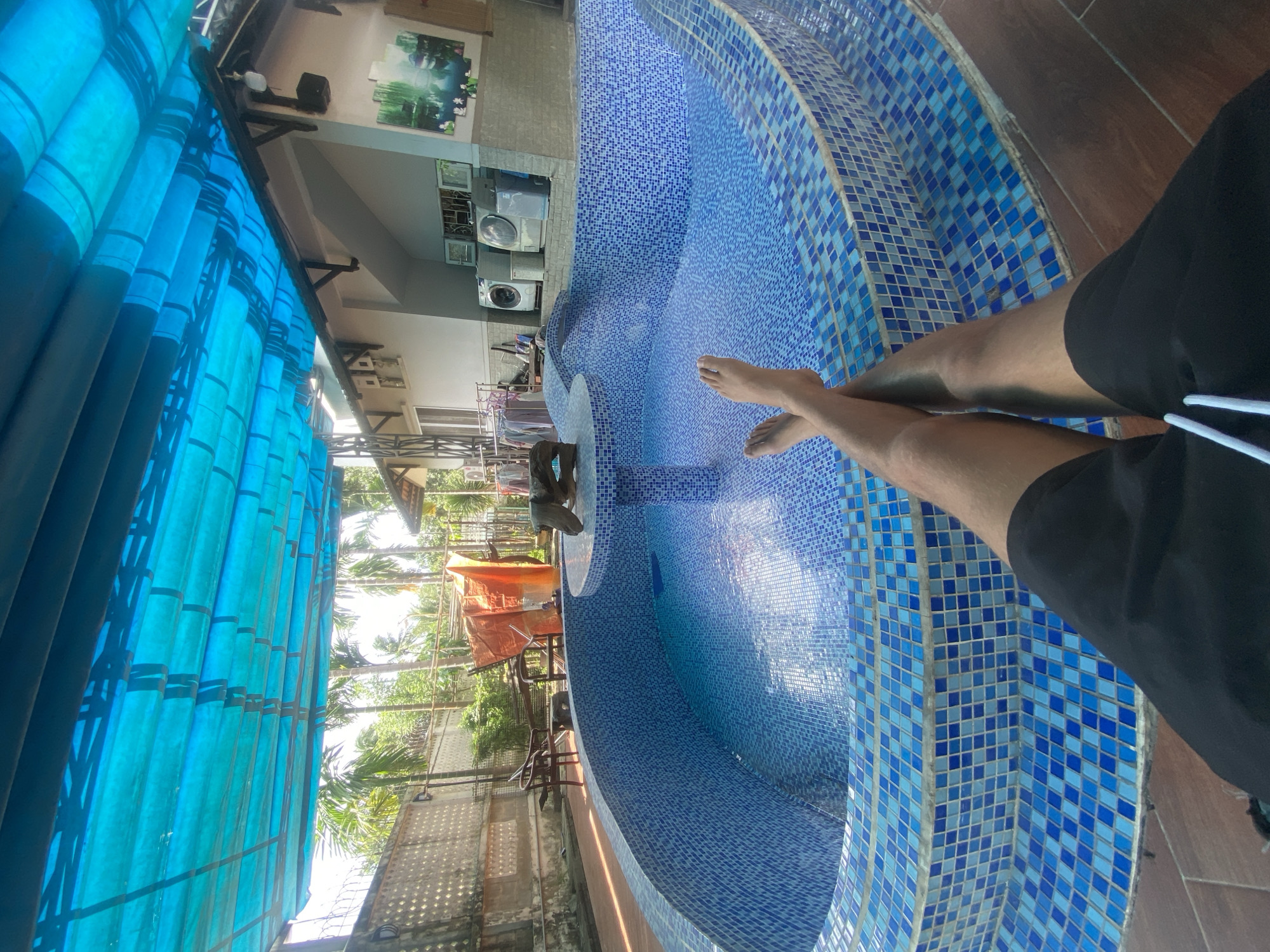 The image depicts a person's legs and feet crossed over each other, resting by the edge of a tiled swimming pool. The pool is kidney-shaped and covered in blue and lighter blue square tiles. There is a central structure in the pool that appears to be a small table or fountain with a decorative element on top. The area surrounding the pool is paved with reddish-brown tiles. Above the pool, a blue tarp is stretched out, supported by a metal structure, providing shade. Beyond the pool, palm trees and lush green foliage are visible, suggesting a tropical or subtropical environment. A building with white walls and a sloped roof is situated behind the pool, and several appliances resembling washing machines are visible on an outdoor surface near the building. The overall scene suggests a private residence or resort in a warm climate. The location is identified as the city of Thuận An, Vietnam.