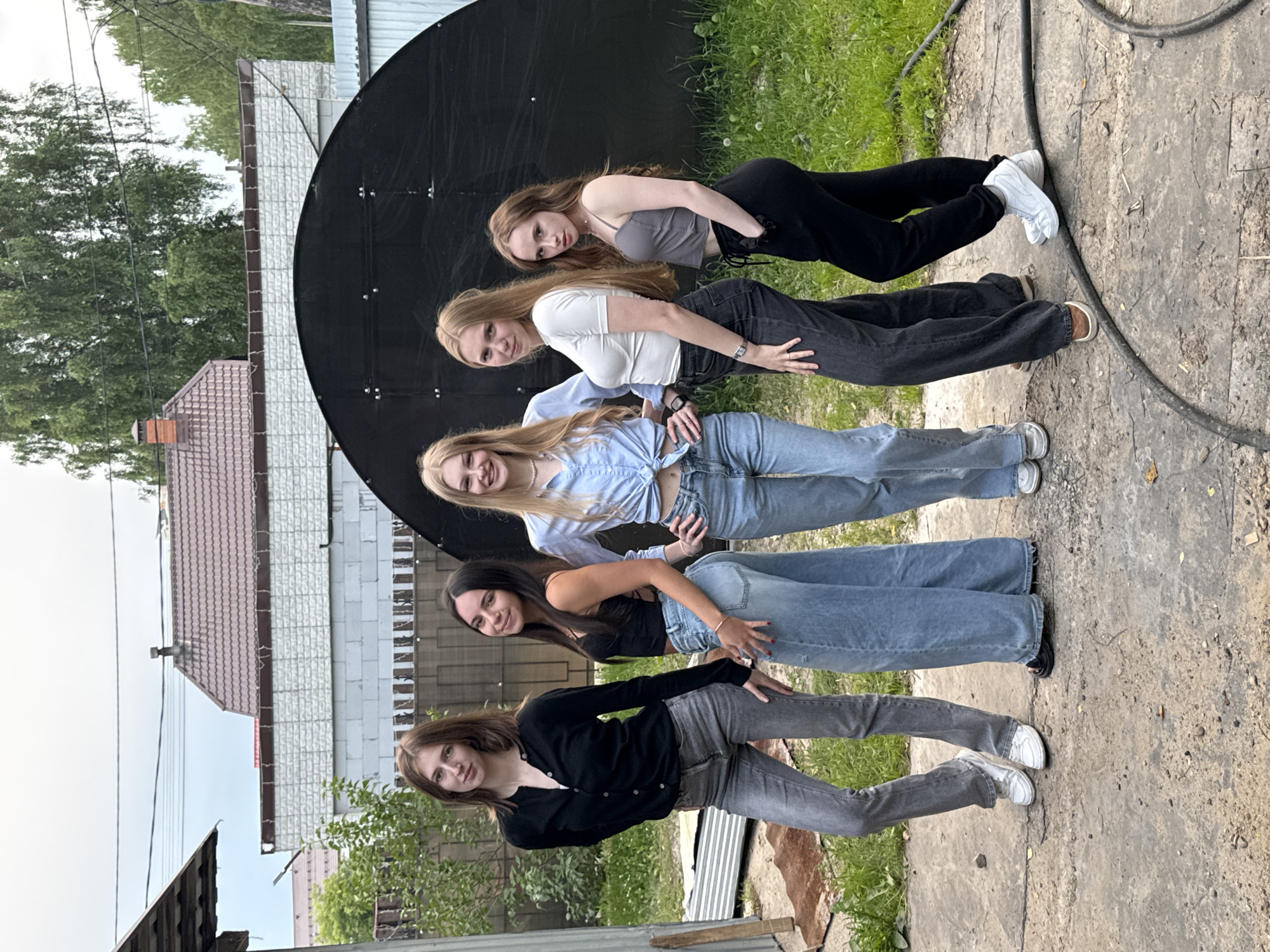 The image shows a group of five young women posing outdoors. They are arranged in a vertical line, with their bodies leaning against a large, black circular object, possibly a satellite dish. The women are dressed in casual, modern clothing, including jeans, crop tops, and cardigans. Some are smiling, and they appear relaxed and friendly. The setting is outdoors, with grass and a concrete path visible. In the background, there are buildings with tiled roofs and white brick exteriors, suggesting a residential area. Trees are also present, with green foliage indicating it is likely springtime or summer. The lighting suggests it is daytime, and the sky appears overcast. There are no visible text elements in the image. The context provided indicates the location is Perm, Russia.