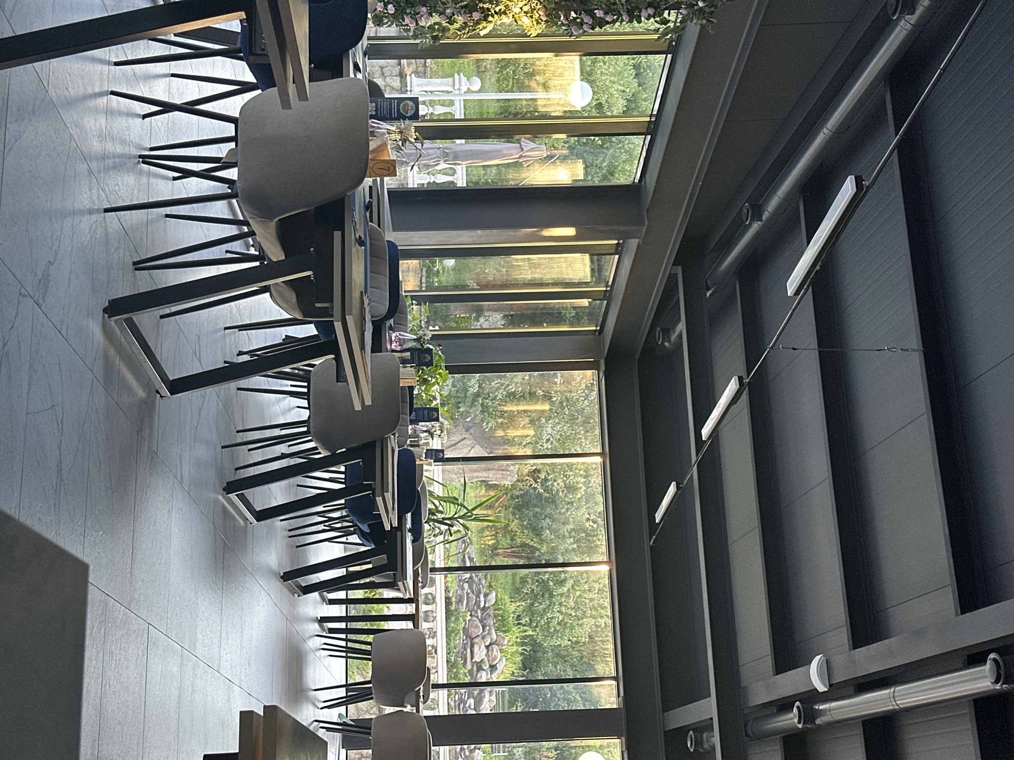 The image depicts the interior of a modern restaurant or cafe with large windows overlooking a lush, green outdoor area. The restaurant features contemporary furniture, including sleek tables with dark metal legs and comfortable upholstered chairs in shades of gray and blue. Long, rectangular fluorescent lights are suspended from the dark, industrial-style ceiling, which also has exposed ventilation pipes.
The floor is tiled with large, light gray rectangular tiles. On the tables, there are place settings with cutlery and what appear to be menus. Decorative elements include small potted plants and floral arrangements, one of which, a vertical cascade of pink flowers, is visible on the left side of the image.
Through the expansive windows, a sunlit garden or park is visible, with trees, stones, and a white balustrade. A closed umbrella and a lamppost are also noticeable in the outdoor scene. The lighting suggests it is either late afternoon or early morning, with a warm, golden hue illuminating the outdoor landscape. There are no people visible in the image. The overall impression is one of a sophisticated and tranquil dining environment with a strong connection to nature through its extensive window views.