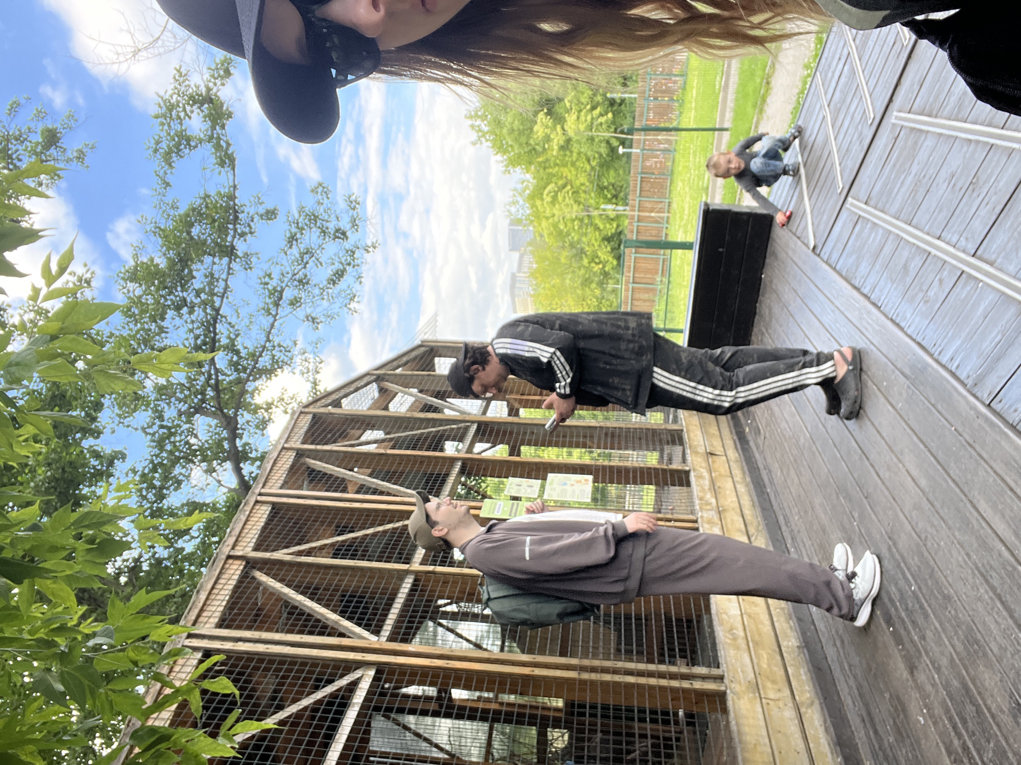 The media file depicts an outdoor scene with a group of people. In the foreground, two young men are visible. One, wearing a dark tracksuit with white stripes, appears to be bending down, possibly interacting with something on a wooden platform. The other, in a light brown tracksuit and a cap, is standing with a backpack and looking upwards. A young child, dressed in jeans and a gray shirt, is crouching on the wooden platform further away, holding a red object.
The setting is an outdoor area with a large wooden structure, resembling an aviary or a zoo enclosure, with mesh fencing. In the background, there are trees, lush green foliage, and a clear blue sky with some clouds, suggesting it is daytime with fair weather. The wooden platform is composed of planks laid out in a linear pattern.
The orientation of the image is rotated. The overall mood appears casual and relaxed, possibly a visit to a park or an animal sanctuary. No discernible text is visible in the media. The location context is Moscow, Russia.