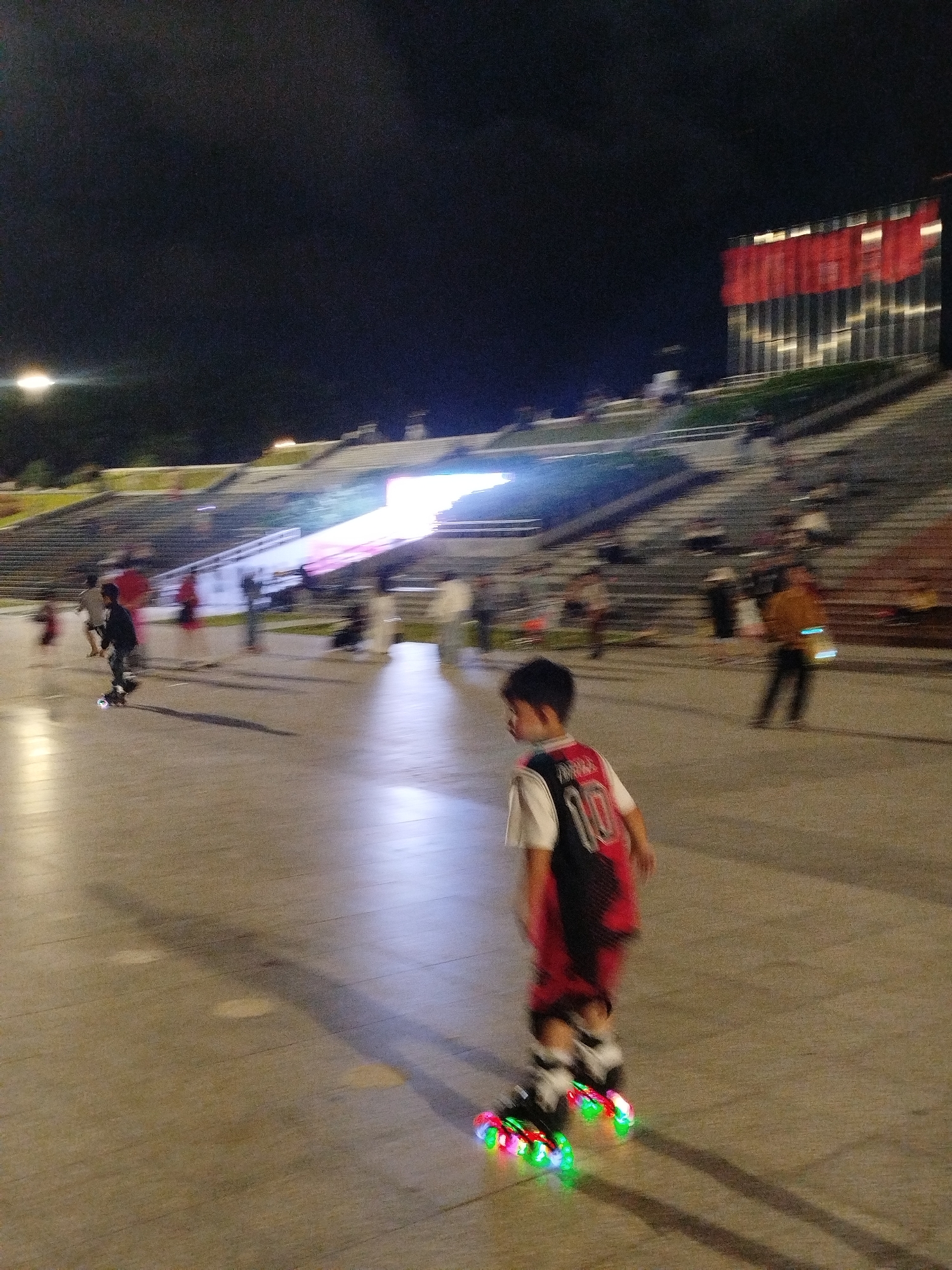 The image shows a group of people in an outdoor stadium or amphitheater setting at night. The primary subject in the foreground is a young boy on roller skates, wearing a red and black jersey with the number 10 visible. His skates have colorful flashing lights. In the background, other individuals are scattered throughout the seating area and on the ground level. The scene is illuminated by artificial lights, including bright lights on a building in the background, which appears to be a modern structure with red banners or panels. The time of day is clearly night. The overall atmosphere appears to be one of leisure and recreation, with people enjoying a public space after dark. The location is identified as Da Lat, Vietnam.