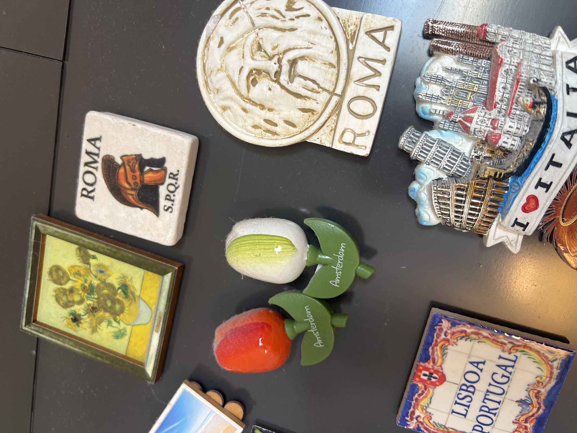 A collection of refrigerator magnets is arranged on a dark, flat surface. The subjects are decorative objects, primarily souvenirs from different locations.
One magnet is a rectangular tile with the word "ROMA" in large letters and an image of a Roman helmet and the inscription "S.P.Q.R." Another magnet features a circular relief with the word "ROMA" and a depiction of what appears to be a Roman profile. A larger magnet depicts an artistic rendition of various Italian landmarks, including the Leaning Tower of Pisa, with "ITALIA" written in prominent letters.
There are also two tulip-shaped magnets with green stems and leaves, one red and fuzzy and the other white with green stripes. Both have the word "Amsterdam" written on the leaves. Adjacent to these is a miniature framed print that resembles Vincent van Gogh's "Sunflowers."
Another magnet is a square tile adorned with traditional Portuguese tile patterns and the words "LISBOA PORTUGAL."
The lighting suggests an indoor setting, possibly a kitchen counter or refrigerator door. There are no people or animals visible in the image. The arrangement of the magnets does not indicate a specific event or activity, but rather a display of collected items. The surface they are on is plain and dark, allowing the colorful magnets to stand out.