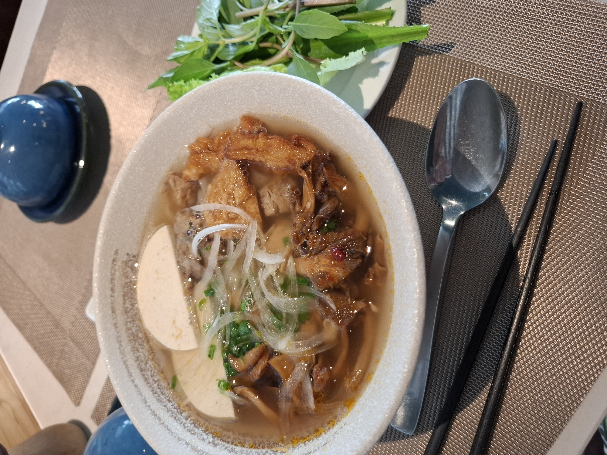 A close-up, high-angle shot shows a bowl of Vietnamese pho on a textured placemat. The pho contains broth, noodles, sliced white meat, green onions, and thinly sliced white onions. To the right of the bowl is a small plate with a bunch of fresh green herbs. A metal spoon and two black chopsticks rest on the placemat next to the bowl. In the background, a small, dark blue, rounded object sits on a dark blue saucer. The scene is lit by natural light, casting shadows on the placemat.