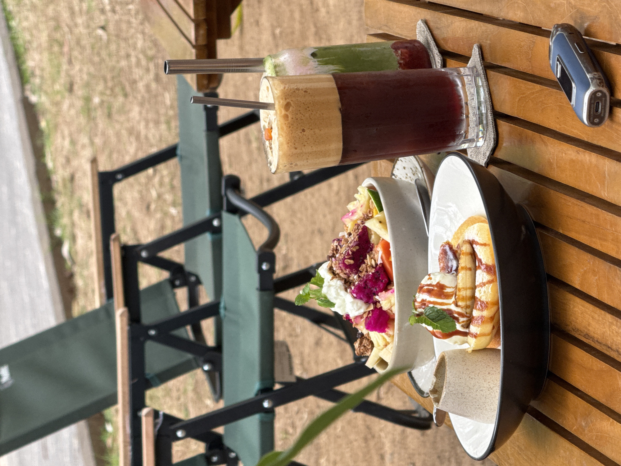 A medium shot captures a wooden table laden with food and drinks, set outdoors in what appears to be a cafe or restaurant in Kuta Selatan, Indonesia. Two folding green camping chairs are visible in the background, suggesting an outdoor seating area.
On the table, a dark-colored beverage with a foamy top sits in a tall glass, accompanied by a green layered drink in a bottle. In front of the drinks, a bowl holds a colorful fruit salad topped with granola and what looks like yogurt or cream. To the left, a small pitcher contains a lighter-colored liquid. Closer to the viewer, a black-rimmed white bowl displays a stack of pancakes topped with a scoop of ice cream, drizzles of syrup, and a sprig of mint.
On the right side of the table, a small, blue electronic device is resting on the wooden surface. The overall scene suggests a leisurely meal or break in a tropical or semi-tropical environment.