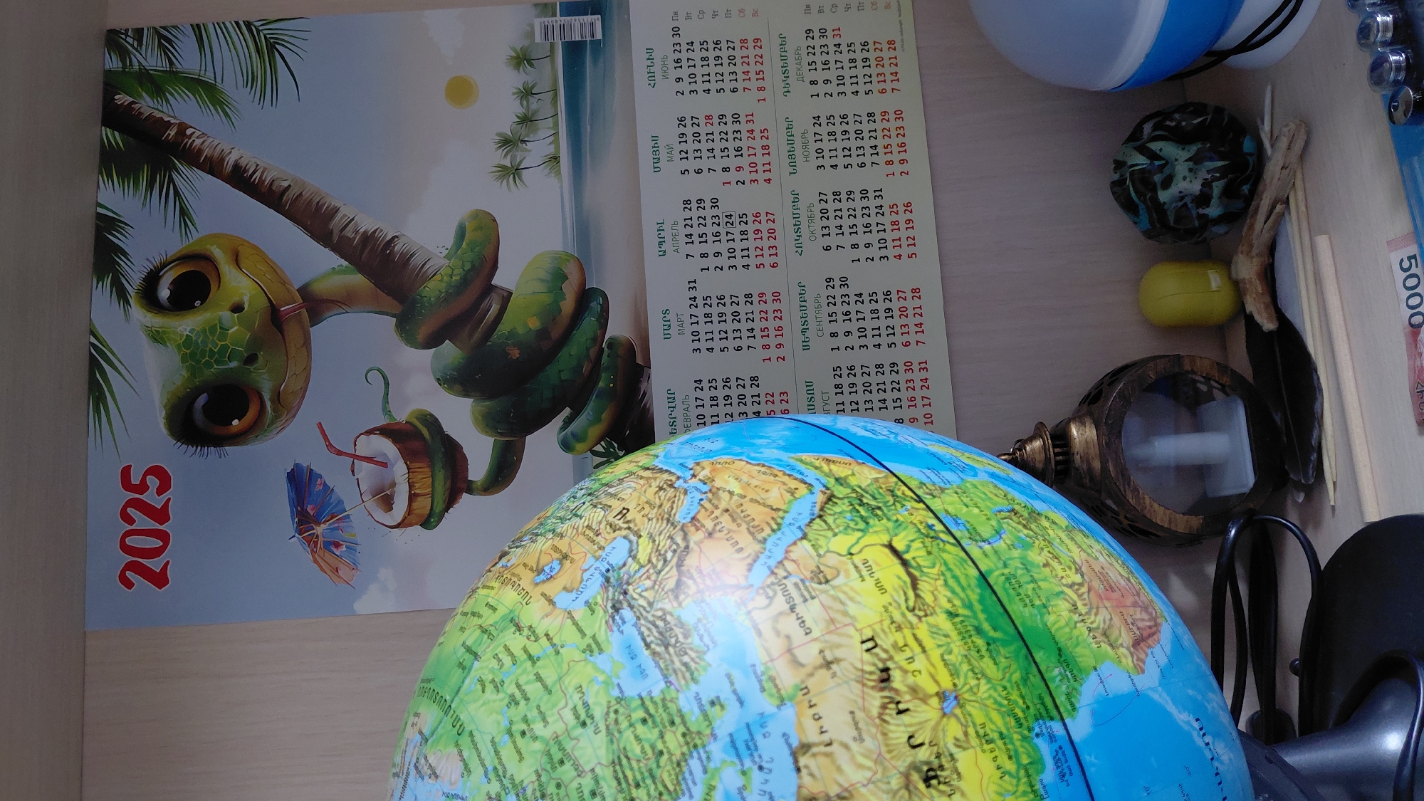 The image displays a corner of a room with various objects on a shelf. A large globe, showing parts of Europe, Africa, and the Middle East, is prominently placed on the left. To its right, a calendar for the year 2025 is visible, featuring a cartoon illustration of a snake on a palm tree, holding a coconut drink with a small umbrella. The calendar has sections with dates in Armenian and Russian. On the shelf below, there's a decorative lantern, a yellow object, a dark, textured sphere, a feather, and some thin wooden sticks. A portion of a blue and white lamp is seen on the far right. The background appears to be a light-colored wall or shelf. There are no people in the image. The presence of Armenian text on the calendar strongly suggests a connection to Armenia, and specifically to the city of Abovyan, given the context provided. The arrangement of objects suggests a personal collection or display.