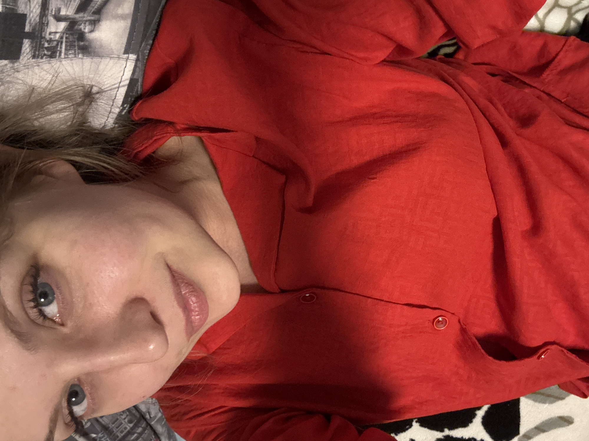 A selfie-style photograph shows a fair-skinned woman with blonde hair and blue eyes wearing a red button-down shirt. She is lying down, facing the camera with a slight smile. To her left is a pillow with a black and white image that appears to depict a cityscape. The background also includes a black and white patterned surface. The photograph does not offer any information to determine if the location is in Izhevsk, Russia.