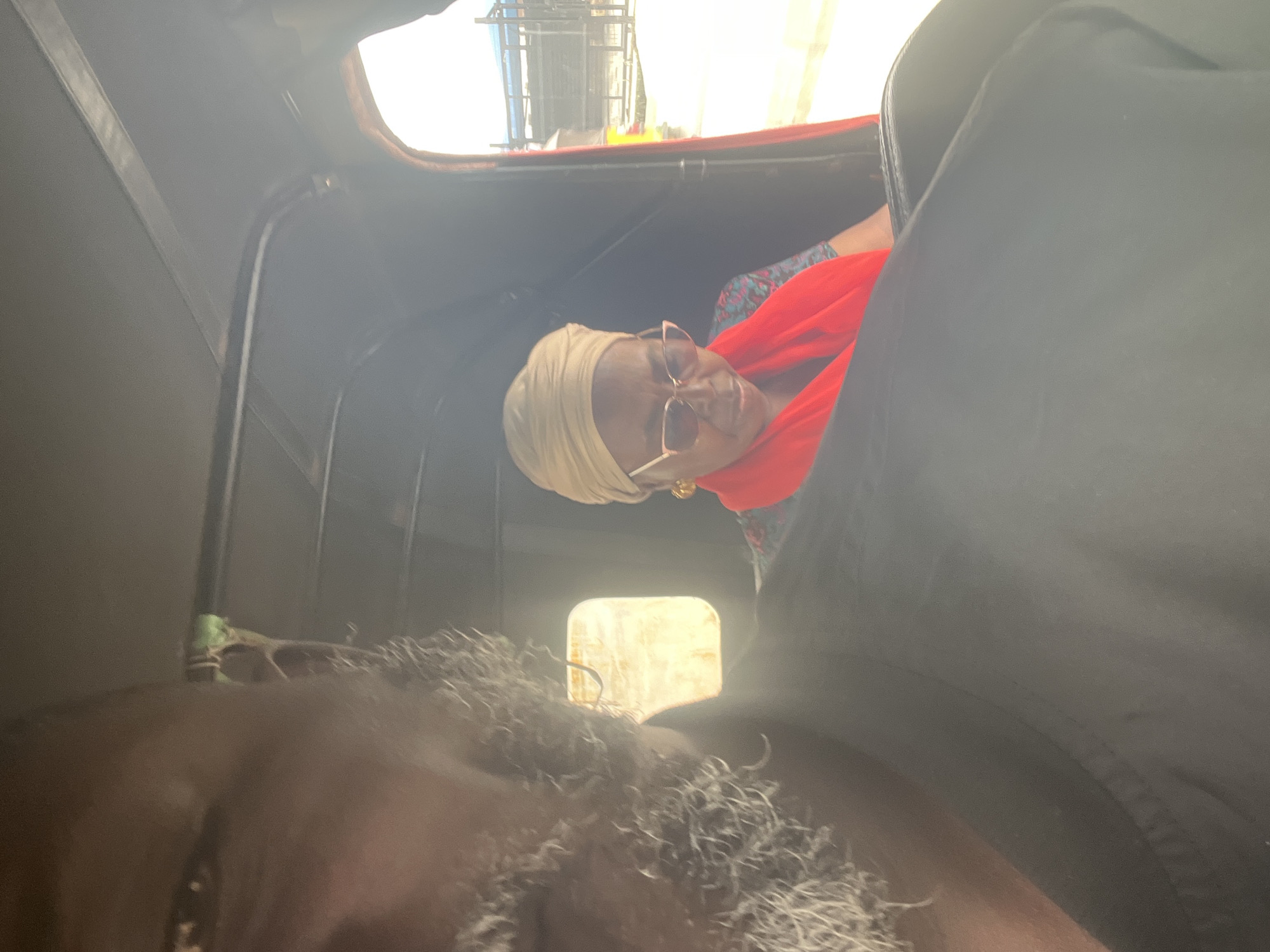 In Ado, Nigeria, two individuals are seated in what appears to be the passenger compartment of a three-wheeled vehicle, likely an auto-rickshaw, possibly in transit. The person in the foreground, whose face is largely out of focus and fills the left side of the frame, has a beard and is wearing a black top. To the right, a woman is seated, wearing a beige headscarf, sunglasses, and a vibrant red scarf over a patterned blue and pink outfit. She is looking down. The interior of the vehicle is dimly lit, with natural light entering from a window or the open side. The background shows a bright, outdoor environment with structures and a vehicle visible, suggesting they are on a street. The image is taken from within the vehicle, looking towards the passengers and the exterior.