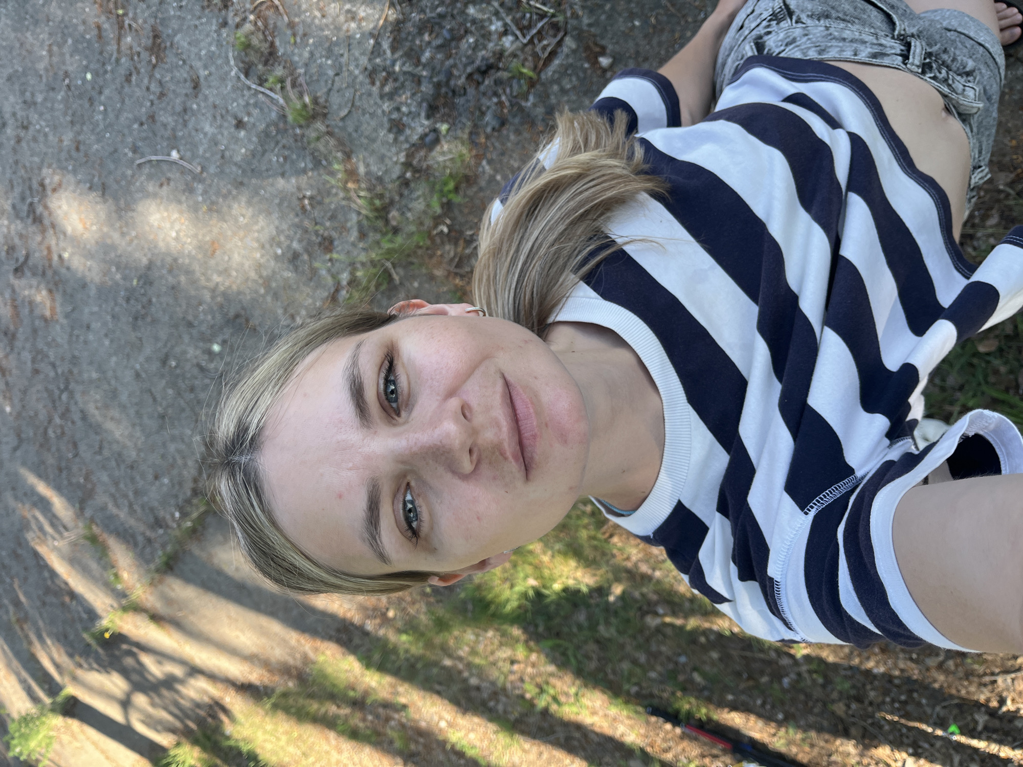 A woman is lying on the ground, taking a selfie. She is wearing a striped shirt and denim shorts. The ground is covered in grass and leaves. A concrete path is to the left. The location is not discernible as Chaikovsky, Russia.