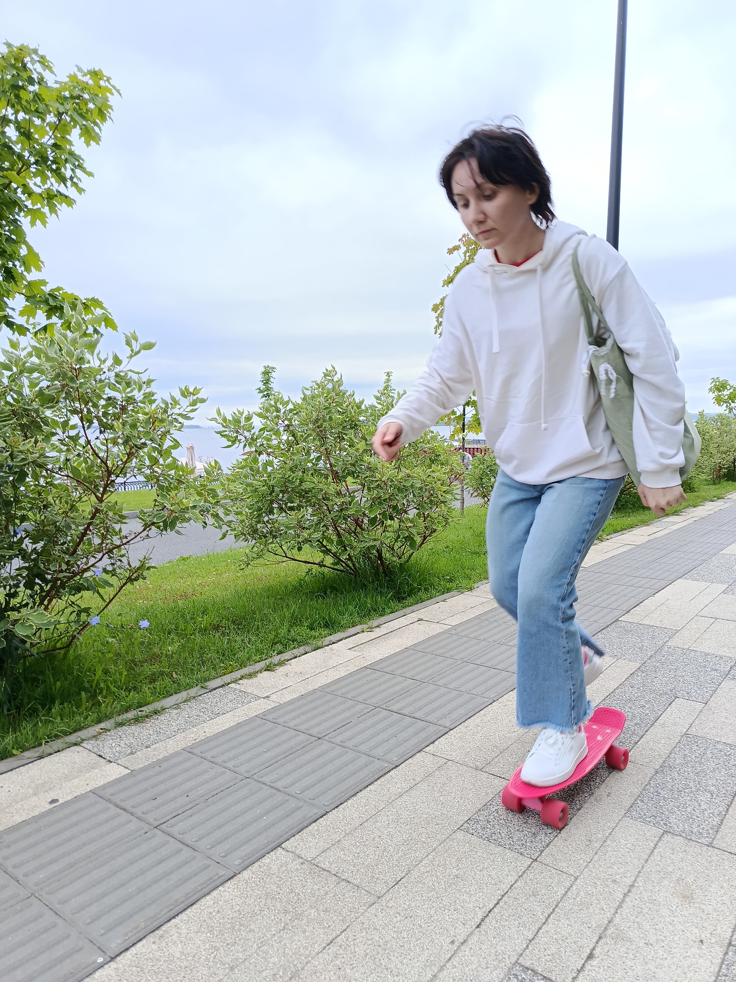 A young woman with dark hair is skateboarding on a sidewalk. She is wearing a white hoodie, blue jeans, and white sneakers. She has a green tote bag slung over her shoulder. The skateboard is pink with red wheels. The sidewalk is made of grey and light-colored paving stones, with textured grey tiles marking a tactile paving path. To the left of the sidewalk is a grassy area with green bushes and trees. In the background, there is a body of water with buildings and structures visible on the other side. The sky is overcast.