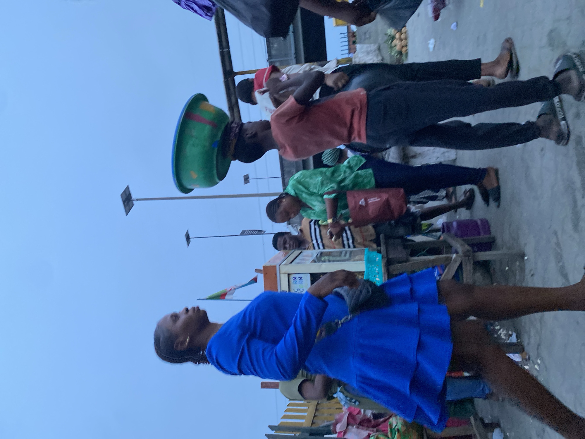 The image captures a bustling street scene in Lekki, Nigeria. A woman in a blue dress walks towards the right of the frame, while other individuals are present near a makeshift stall. One person balances a large green basin on their head. There's a general sense of activity associated with a market or trading area. Streetlights are visible in the background, suggesting a potentially late afternoon or early evening setting.