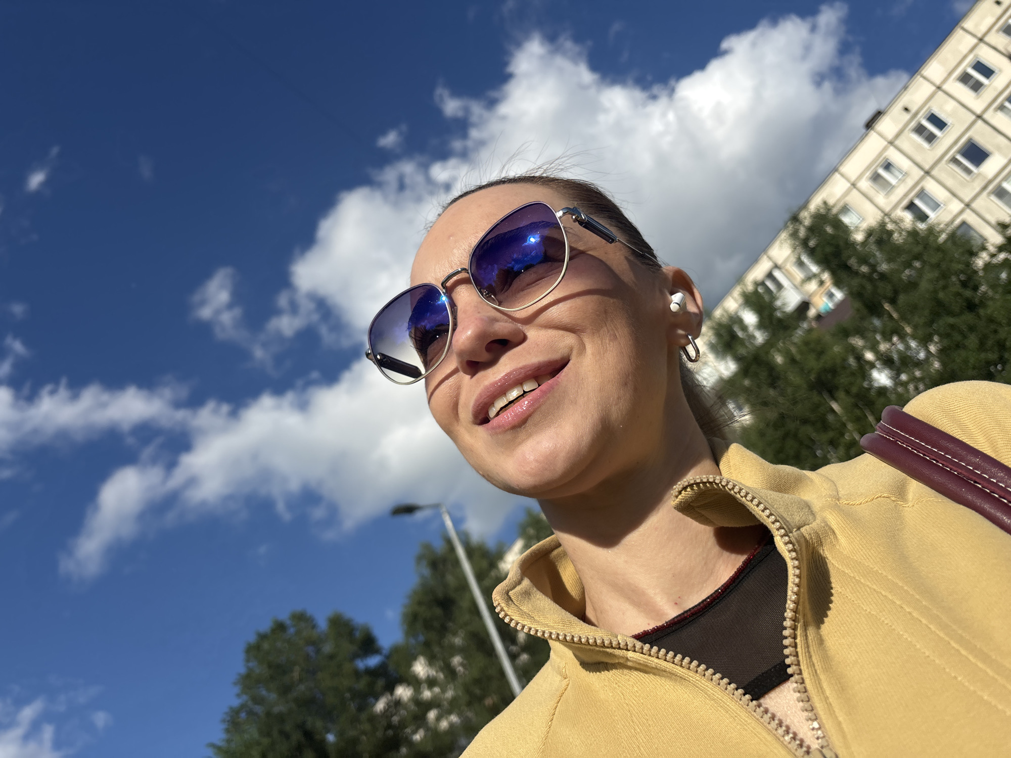 In an outdoor, close-up, low-angle shot, a woman with light skin and dark hair pulled back smiles while looking up and to her left, wearing large, silver-rimmed sunglasses with blue lenses and small white earbuds. She is wearing a yellow zip-up jacket over a dark mesh top. Her left ear is visible, showing a silver hoop earring. In the background, a tall, cream-colored apartment building with many windows dominates the right side of the frame. Trees with dark green foliage are visible in front of the building. The sky is bright blue with scattered white clouds, and a streetlamp is visible on the left side of the frame. The lighting suggests a sunny day.