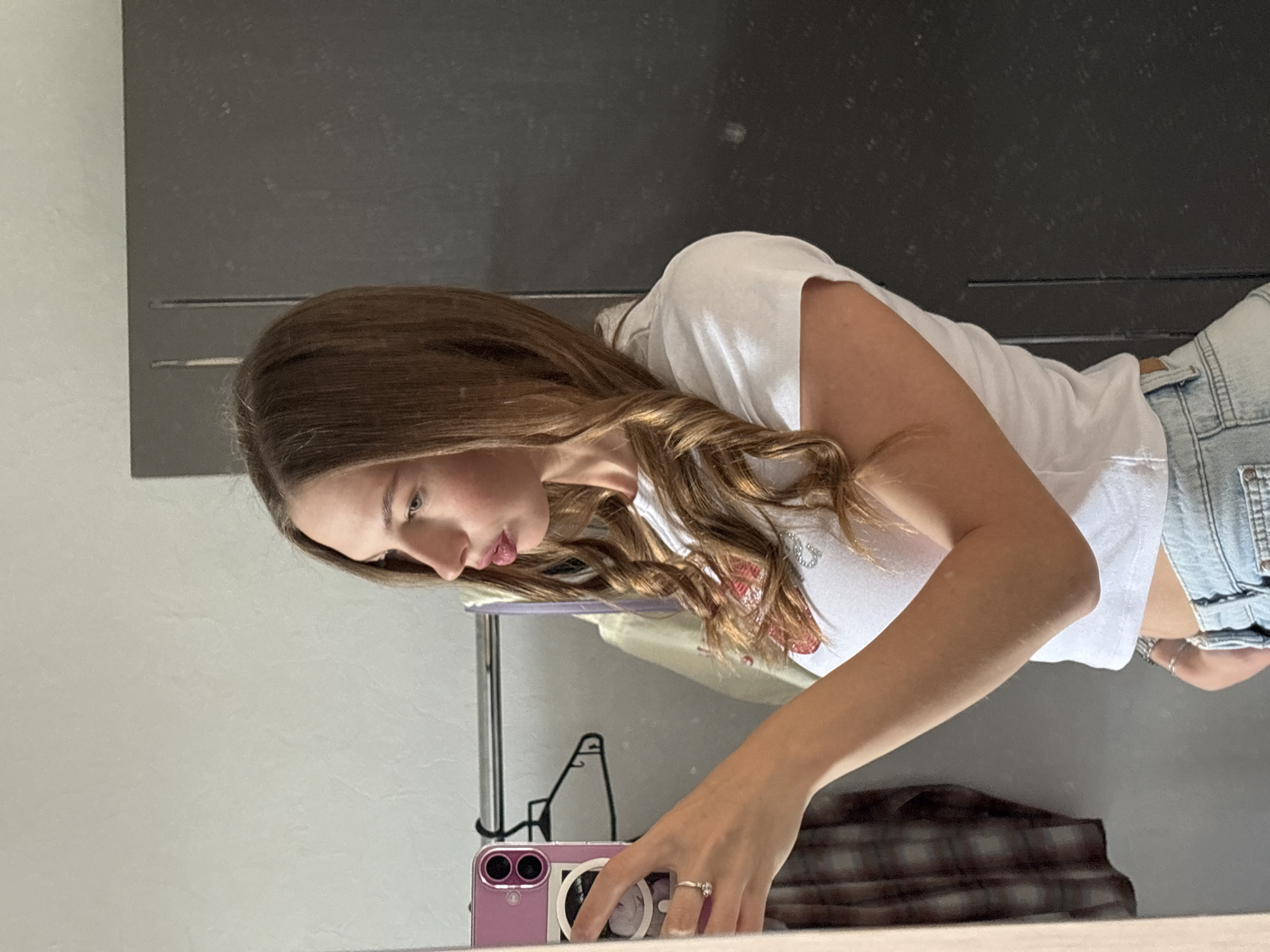 This is a vertical, medium shot of a young woman in her late teens or early twenties, taking a selfie in a mirror. She is positioned in the center of the frame, facing left, with her head tilted down, looking at her phone. The woman has long, wavy brown hair that cascades over her shoulders and down her back. She is wearing a white t-shirt with a red graphic design and denim shorts. Her right hand is visible, holding a pink smartphone and wearing a ring on her ring finger.
The background is a dark, textured surface, possibly a cabinet or a wall, with a lighter, textured wall to the left. A plaid fabric is visible in the reflection in the lower right portion of the image, suggesting furniture or decor. The lighting suggests it is daytime, likely indoors, as there are no strong shadows or indications of natural light sources. The setting appears to be a bedroom or a dressing area. The woman's expression is neutral, focused on taking the photo. No visible text is present in the image.