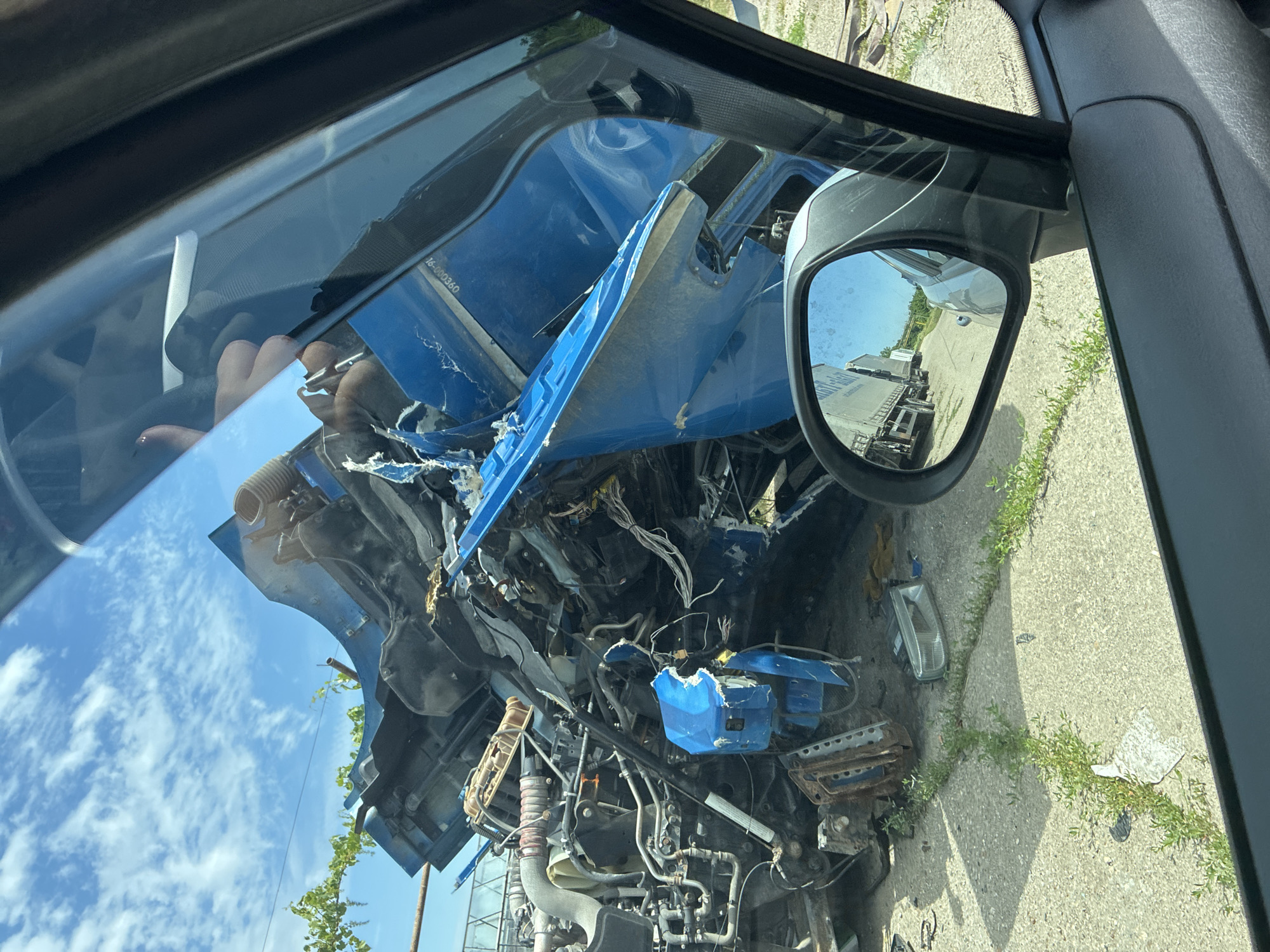 In an outdoor, daytime scene, a severely damaged blue semi-truck is the primary subject. The truck's front end is almost entirely destroyed, with the hood ripped open and various mechanical components exposed, including an engine, hoses, and wiring. Debris is scattered around the truck, with a detached headlight visible on the ground. The truck is parked on a concrete surface with weeds growing through cracks. A side-view mirror of another vehicle, likely a car, is in the foreground, reflecting a line of other trucks on a road. The sky is blue with scattered white clouds. The location is described as Unknown, Unknown.