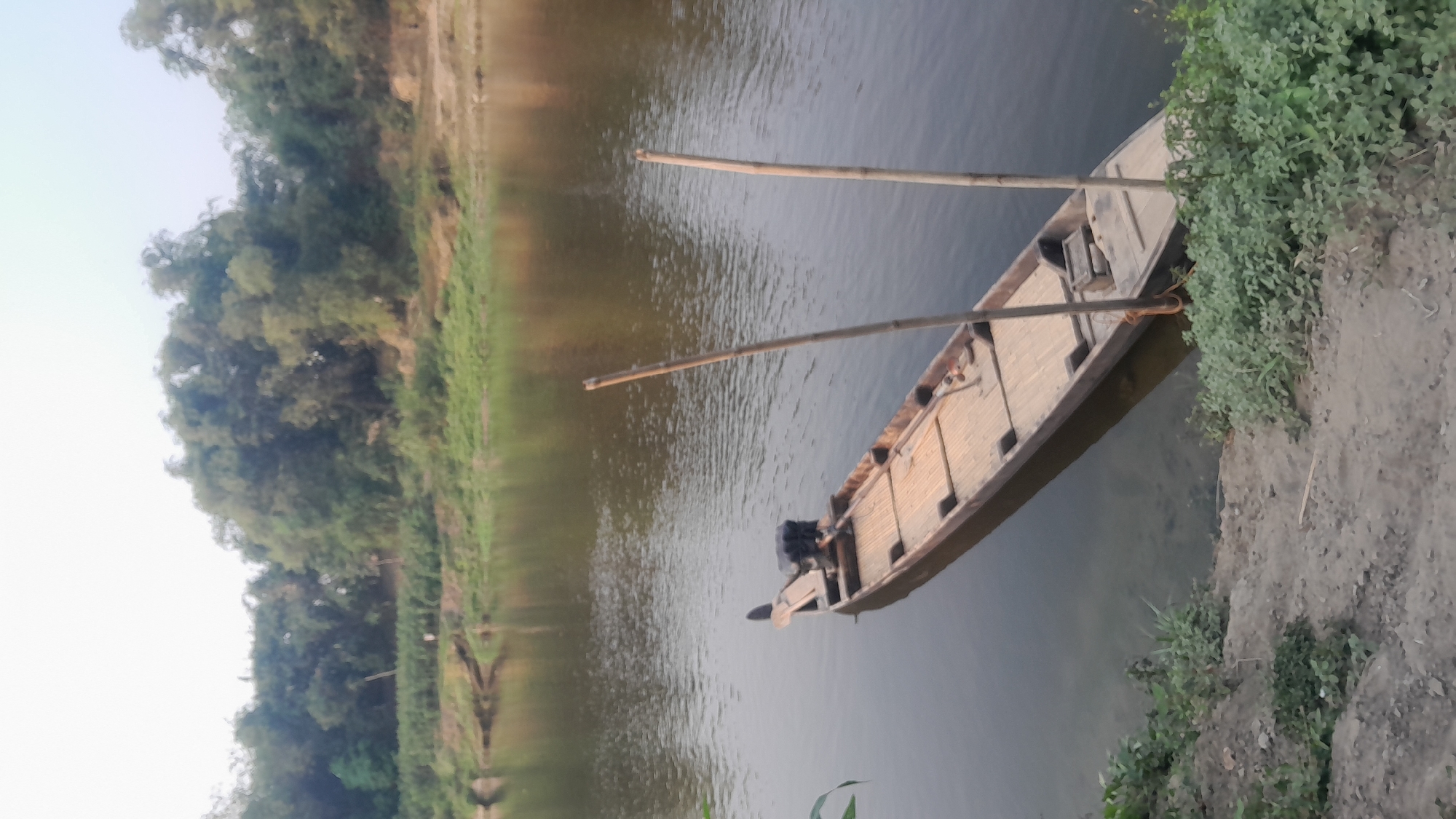 A long, narrow wooden boat with a bamboo floor is moored in shallow, murky water near the bank of a river. The boat is equipped with an outboard motor at its stern and two tall bamboo poles stand upright in its center. The river is calm and reflects the overcast sky and the dense, green foliage of trees that line the opposite bank. The bank in the foreground is a mixture of sandy soil and green vegetation. The scene suggests a quiet, natural setting, possibly in a rural area of Sumberarum, Indonesia.