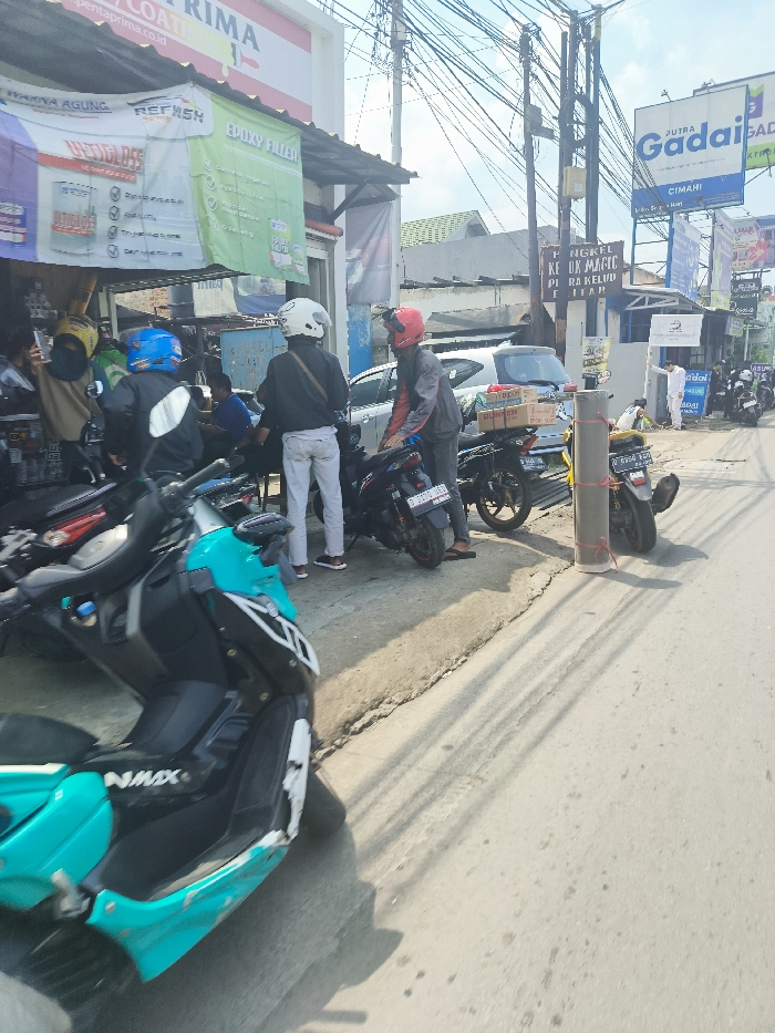 The scene is a street in Ngamprah, Indonesia, with a shop on the left side of the image. Several people are present, including four men wearing helmets and one man in a white shirt. The man in the white shirt is standing near a sign. There are five motorcycles parked along the street. The motorcycles are all facing the same direction, and the men are standing near the motorcycles. The shop has various signs and a blue awning. The street is paved and there is a sidewalk on the right side of the image. The sky is clear and blue with white clouds. There are electrical wires strung above the street. The location is the city of Ngamprah in Indonesia, based on the latitude -6.868 and longitude 107.522.