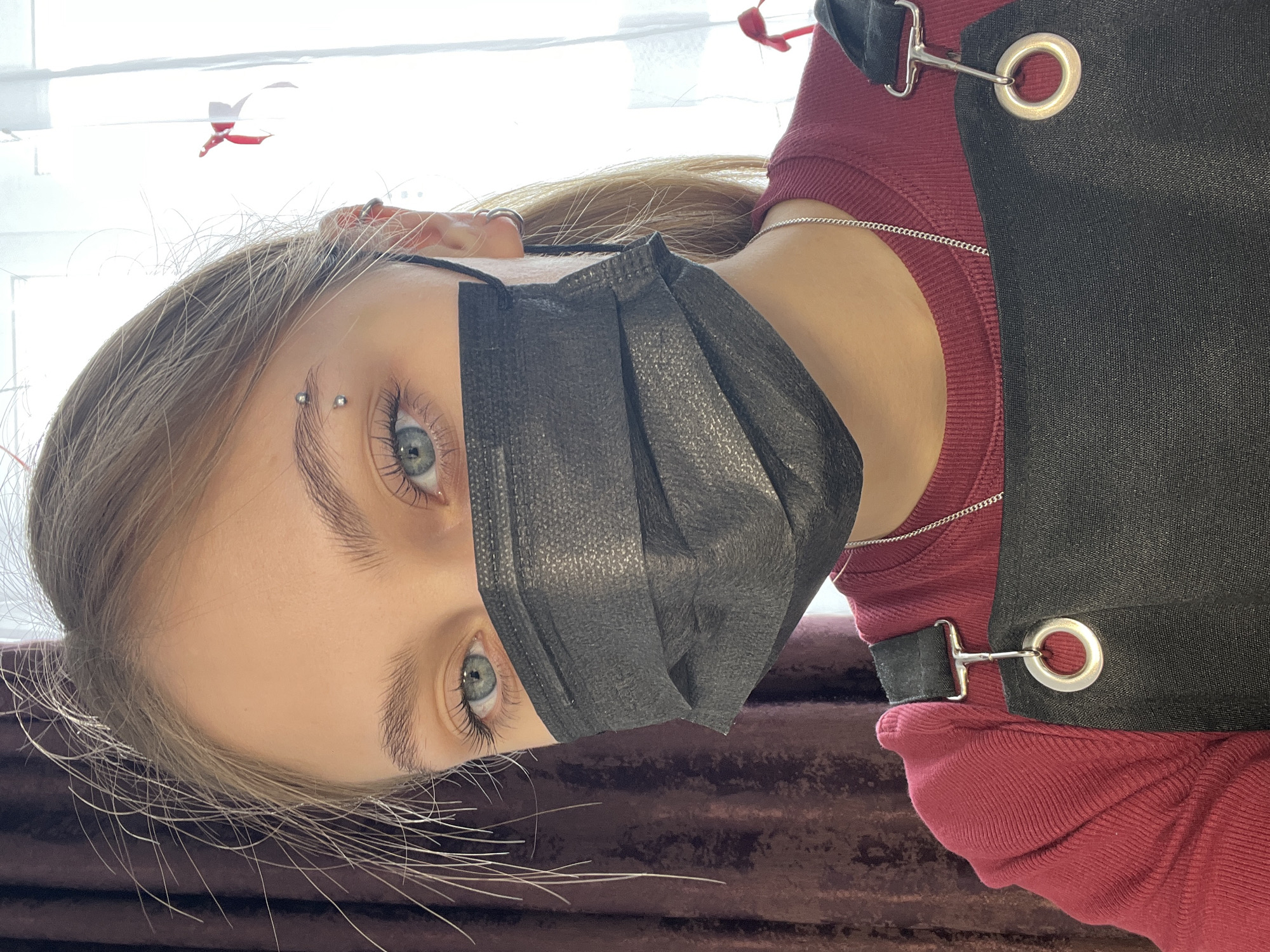 A young, fair-skinned individual with blonde hair is positioned in a close-up shot, wearing a black face mask and a maroon long-sleeved shirt. The individual has an eyebrow piercing. A black apron with silver hardware is worn over the shirt. The background features a window with a red ornament or decoration visible near the top left corner. The image appears to be taken indoors. There is no evidence to suggest a location in the city of Фоки, Russia.