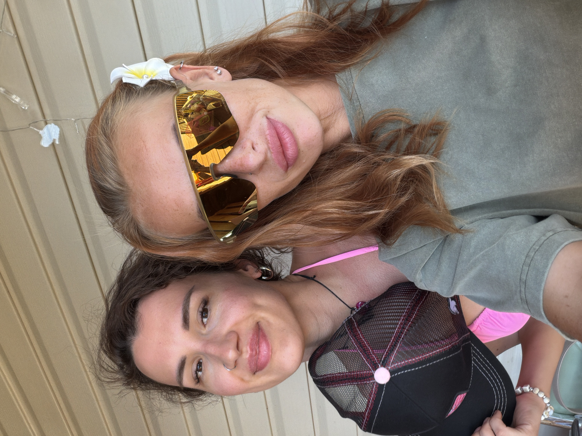 Two women are posing for a selfie outdoors. The woman on the left has dark brown hair, a nose ring, and is wearing a pink bikini top and a black baseball cap with pink accents. The woman on the right has reddish-blonde hair, is wearing large gold-tinted sunglasses with a flower tucked behind her ear, and a green t-shirt. The background appears to be a light-colored, horizontally-paneled wall, possibly on a porch or patio. There are also some blurred string lights in the background. The scene is brightly lit, suggesting it is daytime.