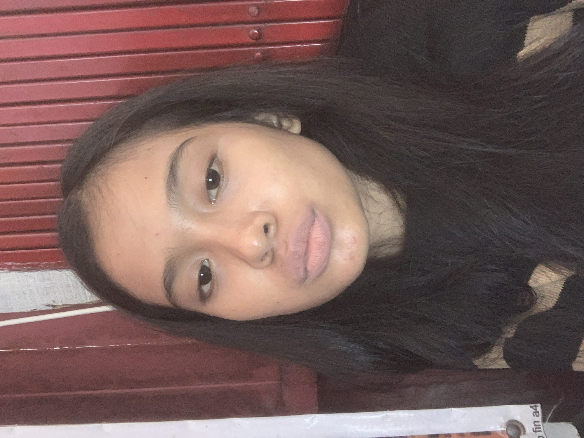 The media file presents a close-up portrait of a young person, likely female, with long, dark, straight hair. The image is rotated, showing the subject's head tilted to the side as they look directly into the camera with a neutral to slightly pouting expression.
The subject's facial features include dark eyes, natural-looking skin with visible pores, and slightly parted lips. They are wearing a dark top, possibly a sweater, which features a distinct beige or light brown textured pattern visible on the right side of the frame.
The background primarily consists of a deep red surface with horizontal grooves, resembling a metal roll-up door or a corrugated wall. This red surface dominates the upper and left parts of the image. At the bottom of the frame, there is a horizontal white surface, possibly a ledge or a sign. In the bottom right corner, partially obscured, the text "fin a4" is visible on this white surface, near what appears to be a metal fastener or clip on a piece of paper or board.
The lighting suggests it is daytime, providing soft and even illumination on the subject's face. The setting appears to be an indoor or sheltered outdoor space, potentially in Sungai Meriam, Indonesia. The visible activity is the subject posing for the photograph.