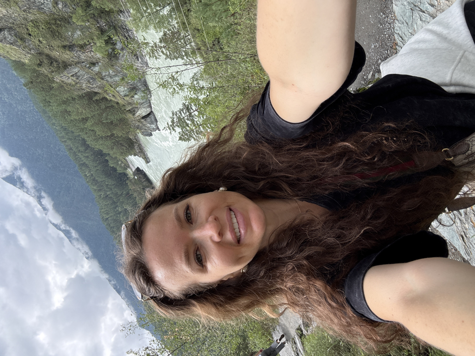 In a selfie taken outdoors, a woman with long, wavy brown hair smiles at the camera. She is wearing a black t-shirt and a light-colored hooded garment. Sunglasses are perched on top of her head. Behind her, a wide, turquoise river flows swiftly through a rocky gorge. Steep, tree-covered slopes rise on either side of the river, and dramatic, cloud-shrouded mountains loom in the background. A second person, whose legs and lower body are visible, is walking away from the camera on a path to the left. The scene appears to be in the mountainous region near Chemal, Russia, likely along the Katun River, a popular tourist destination known for its scenic beauty.