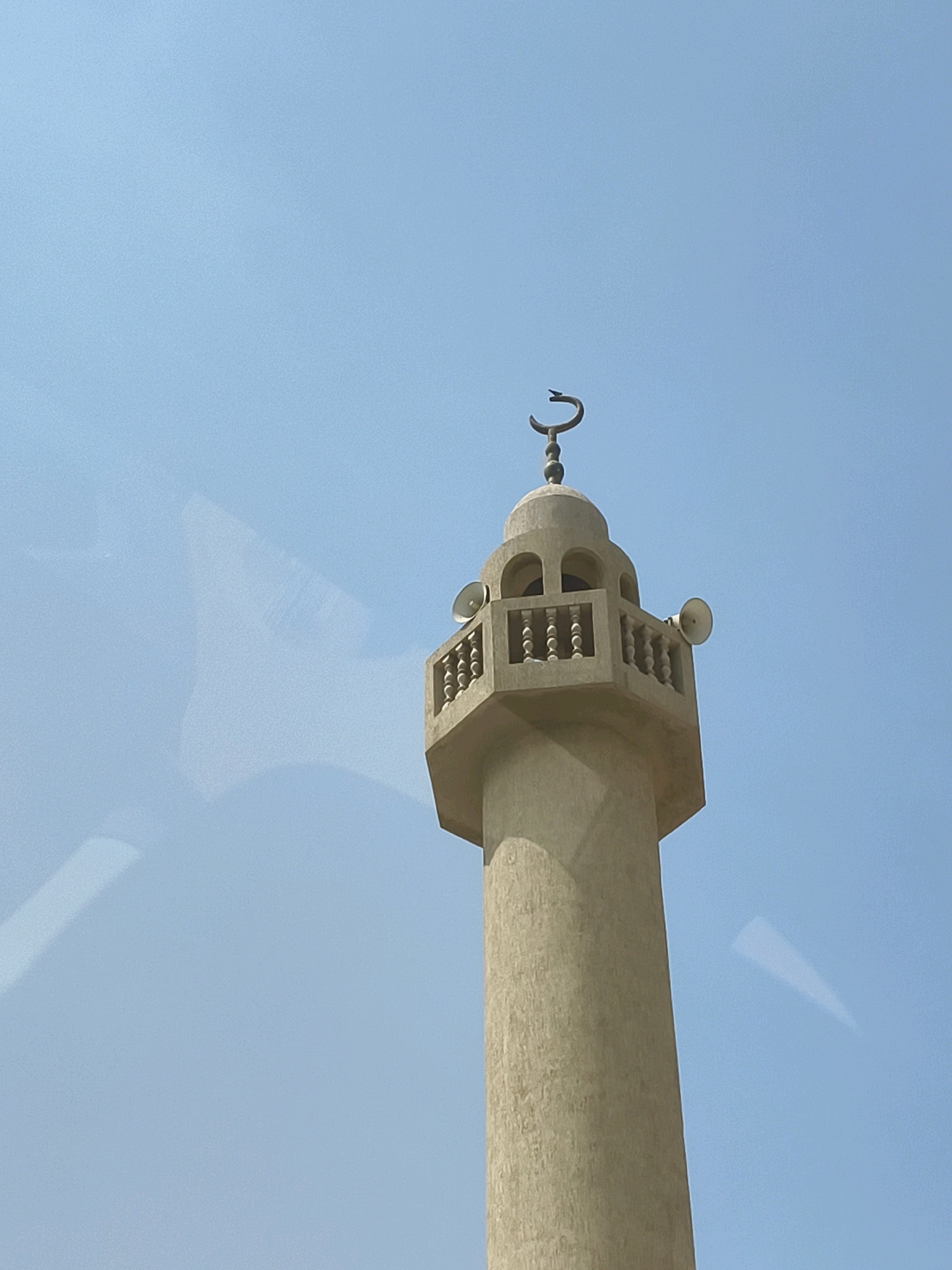 A minaret stands against a clear blue sky. The minaret is made of light-colored concrete and has an octagonal base. It is topped with a dome and a crescent moon symbol. Two loudspeakers are mounted on the sides of the upper section of the minaret. The image appears to have been taken from a slightly elevated perspective, possibly from a vehicle, as evidenced by a reflection on the left side of the frame. The setting is outdoors in a bright, sunny environment. The image does not contain any people or specific landmarks that would identify its location in Dubai, United Arab Emirates.