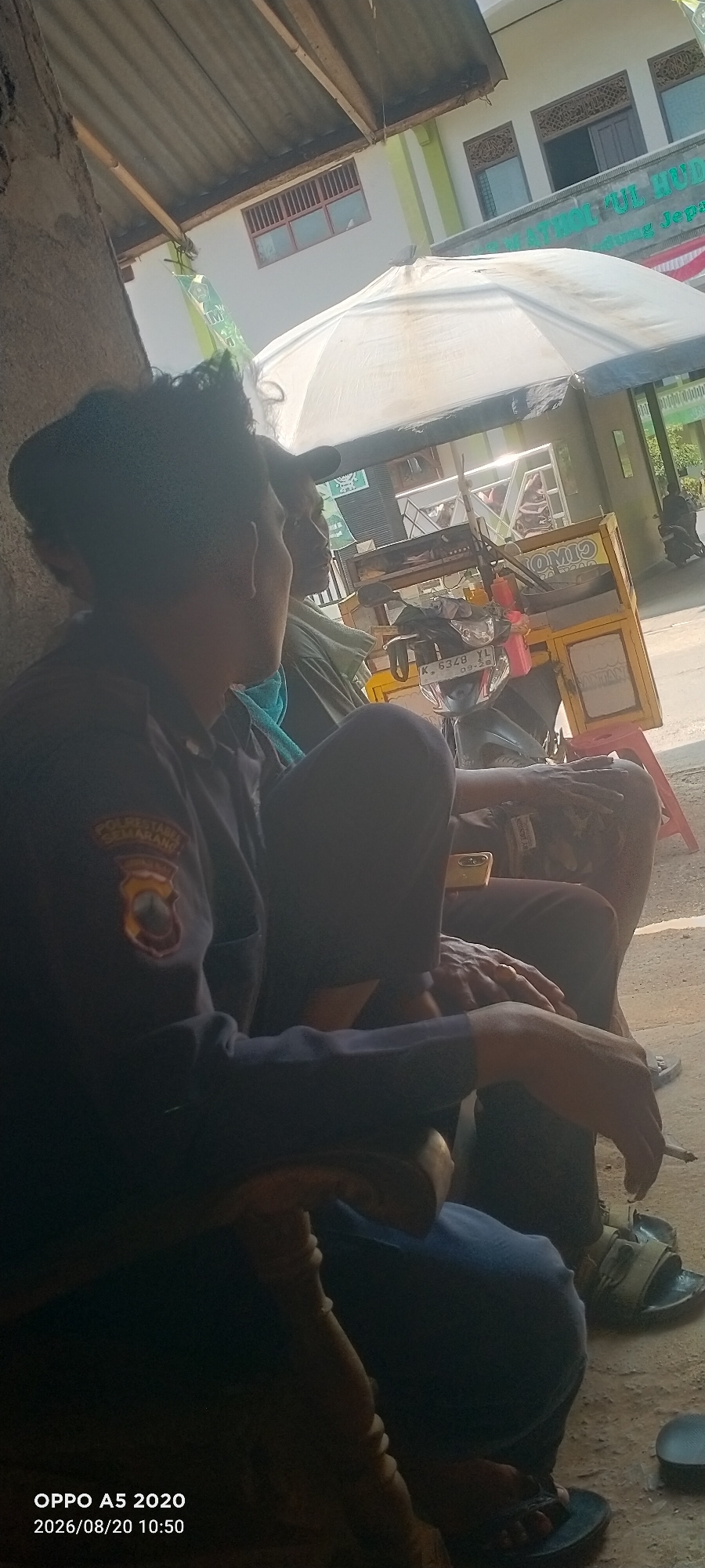 Two individuals are seated outdoors under a thatched roof structure, likely a roadside shelter or pavilion, in Bugel, Indonesia. The person in the foreground, wearing a dark uniform with a patch on the left sleeve, is facing left and appears to be looking at something out of frame. They are wearing dark pants and black sandals. To their right and slightly behind, another person is also seated, wearing a light-colored shirt and dark pants. Their face is partially visible, and they are looking in the same direction as the first person.
In the background, a white umbrella is set up, partially obscuring a mobile food cart with yellow and black signage. The cart is on a motorcycle with a license plate visible that reads "K 6348 YL 09-26". The environment appears to be a street or pathway with a building in the background that has windows with red bars and decorative wooden elements. The ground is dirt or gravel. The lighting suggests it is daytime, with bright sunlight casting shadows. A cigarette is visible in the hand of the person in the foreground, suggesting they are smoking.