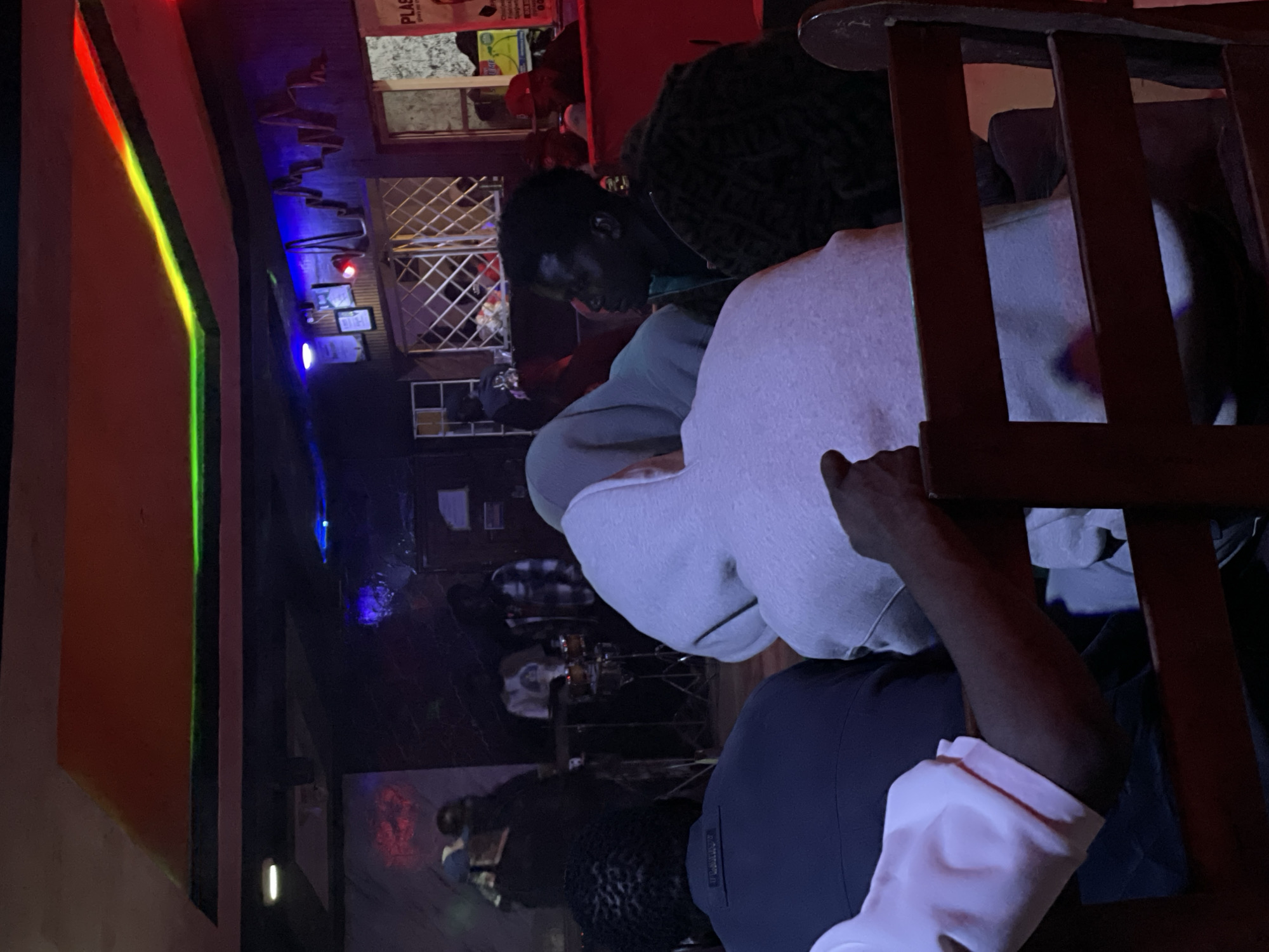 The image depicts an indoor scene at a nightclub or bar in Durumi, Nigeria. The lighting is dim and features a colorful light display on the ceiling with red, green, and blue hues. In the foreground, several individuals are seated at tables, facing away from the camera or in profile. One person in the center is wearing a light gray hooded sweatshirt, obscuring their face. To their left, a person in a dark blue shirt with "FLLRROY" printed on the back is visible. To the right of the hooded figure, a person with dark skin and braided hair is looking to the side, wearing a dark patterned jacket. In the background, a band or group of musicians is performing on a stage. One individual appears to be playing a drum kit, while others are standing near musical equipment. The environment suggests a lively atmosphere with patrons watching the performance. The overall impression is of a social gathering in a dimly lit entertainment venue.
