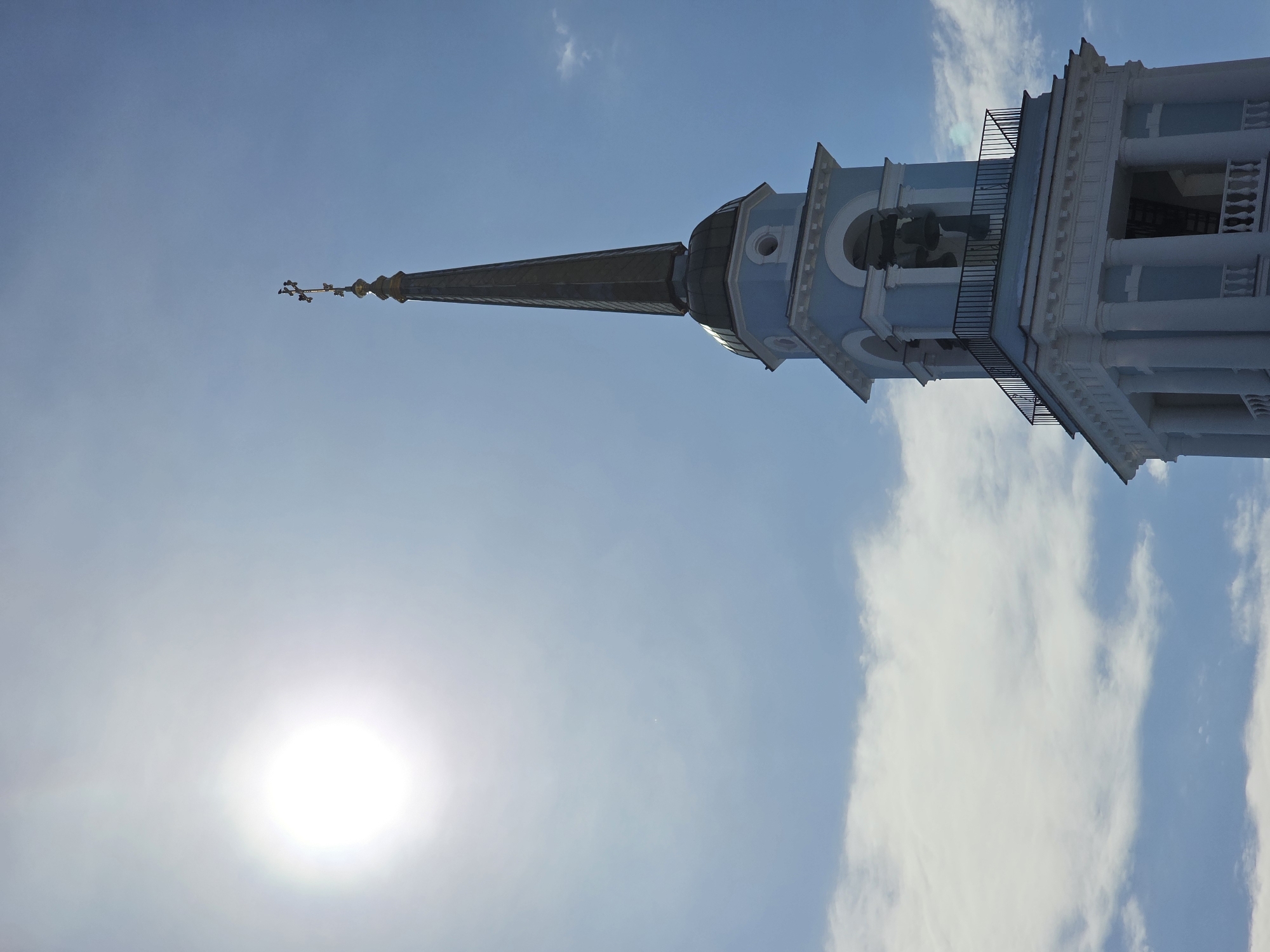 An upward-angled photograph depicts a prominent, multi-tiered bell tower and spire, identified as belonging to a religious edifice, likely the Holy Dormition Cathedral, in Votkinsk, Russia. The tower presents a light blue and white color scheme, featuring white classical columns and a balustrade on its lower visible section. An upper belfry tier contains arched openings revealing dark bells, encircled by a black metal railing. Above this, a dark, domed section supports a tall, slender, dark gray spire. A golden Orthodox cross, characterized by multiple crossbars, crowns the spire. The sun is visible as a bright white disc in the upper-left sky, against a predominantly clear blue sky with scattered white clouds.