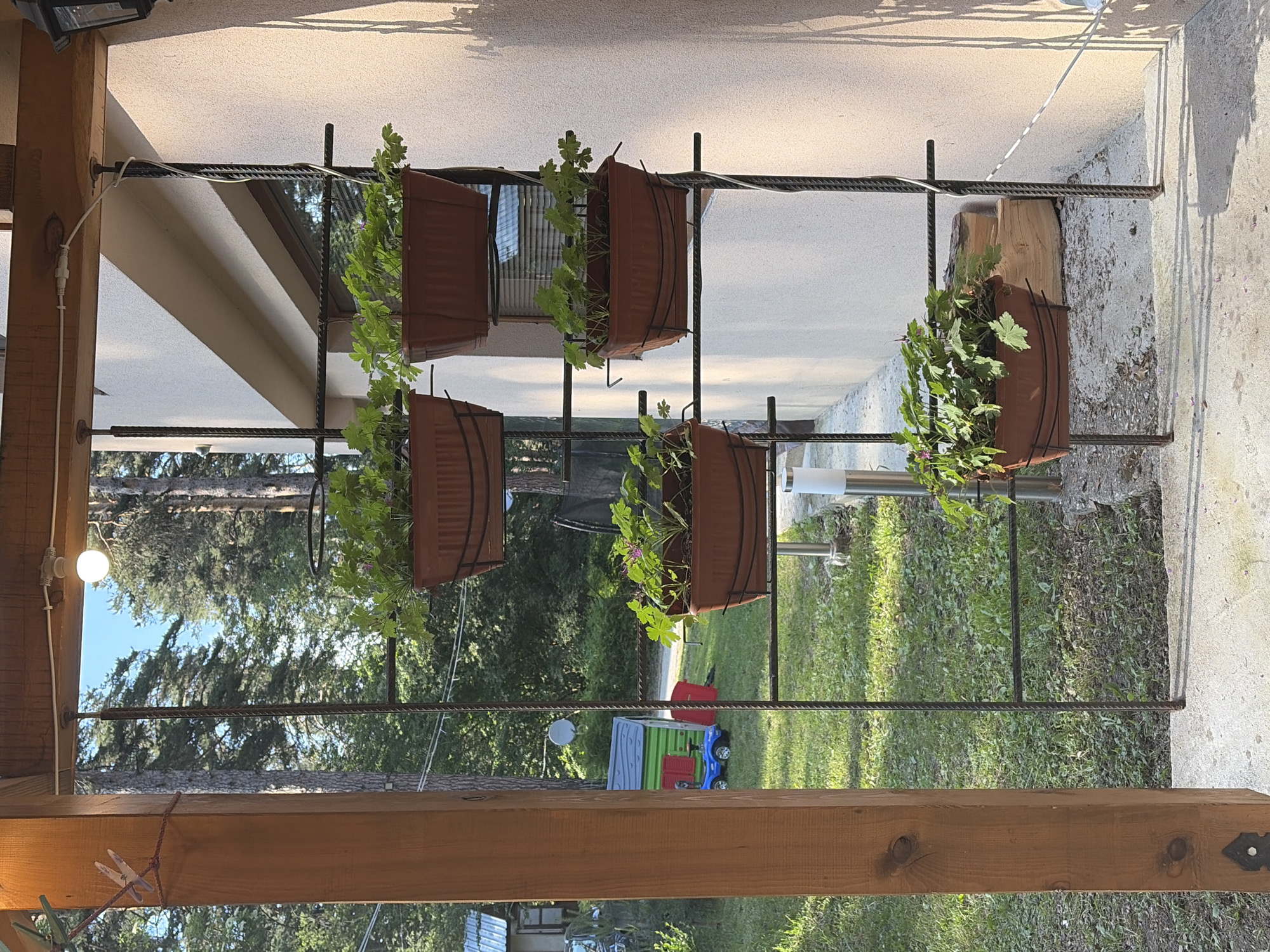 A sunlit outdoor scene features a vertical metal planter structure holding five brown rectangular pots filled with green leafy plants. The planter is attached to a wooden support post on the left, which is anchored to a concrete ground. Behind the planter, a light-colored building wall is visible, with shadows cast by the planter and surrounding vegetation. The background reveals a grassy yard with trees, a small building with a metal roof, a colorful playhouse, and a blue toy car. A single lightbulb hangs from a wire above the planter. The overall environment suggests a residential or garden area in a wooded location.