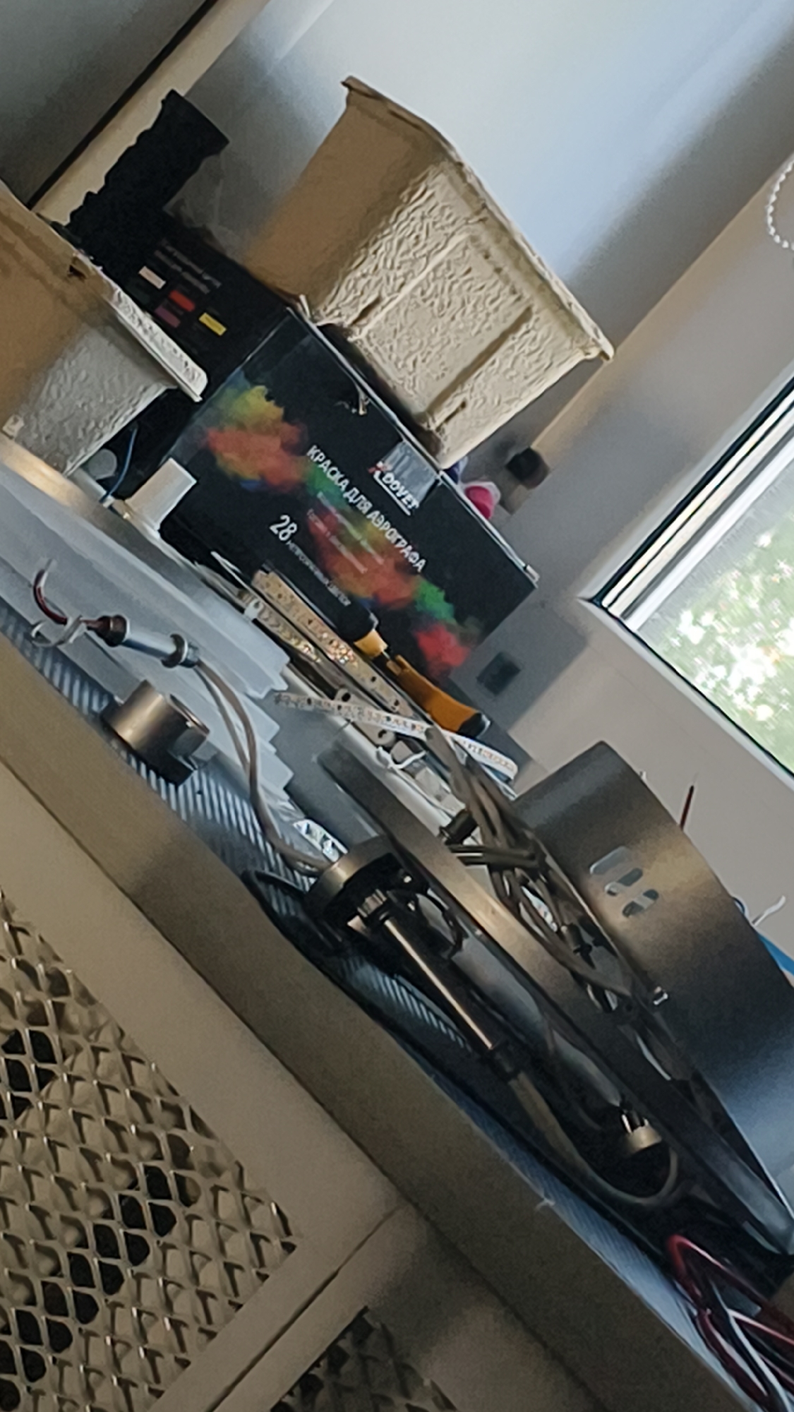 The image shows a collection of items on a shelf or table, possibly in a workshop or storage area. In the foreground, there is a metallic object with a gridded surface. Behind that, a tangle of wires and metal components is visible, suggesting some kind of machinery or electronics. A black box with colorful graphics and text in Cyrillic, identifying it as "КРАСКА ДЛЯ АЭРОГРАФА" (paint for airbrush) and showing the number "28", is prominent. A small yellow object, possibly a tool, is placed on top of this box. To the right, a window with blinds offers a view of greenery outside. The overall impression is one of a cluttered workspace with art supplies and equipment.