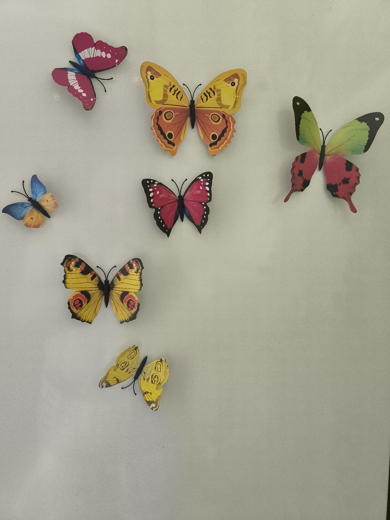 A collection of seven artificial butterflies are attached to a textured, light-colored wall. The butterflies vary in size and color, with designs in shades of pink, yellow, orange, blue, and green. Some butterflies have black antennae. The wall appears to be indoors, with a subtle speckled texture. There are no people or animals visible in the image, nor any indication of the specific location within Rybnitsa, Moldova.