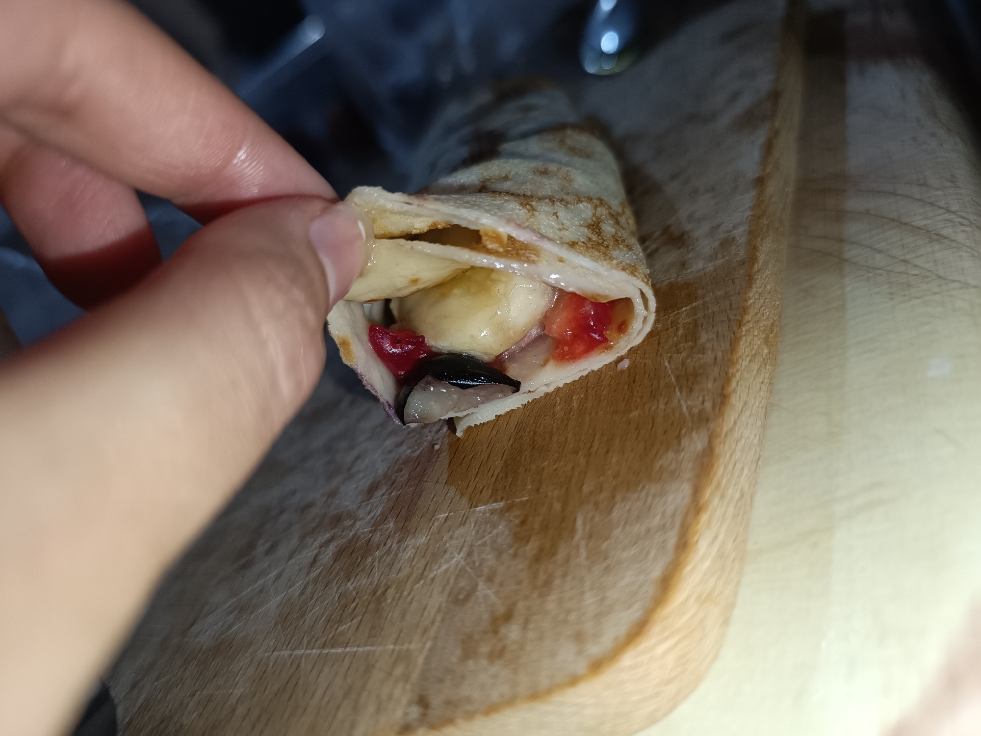 A hand holds a rolled crepe, showing a cross-section of its contents. The crepe is filled with sliced banana, chopped strawberries, and a dark, possibly balsamic glaze. The crepe sits on a wooden cutting board. No location is discernible from the image.